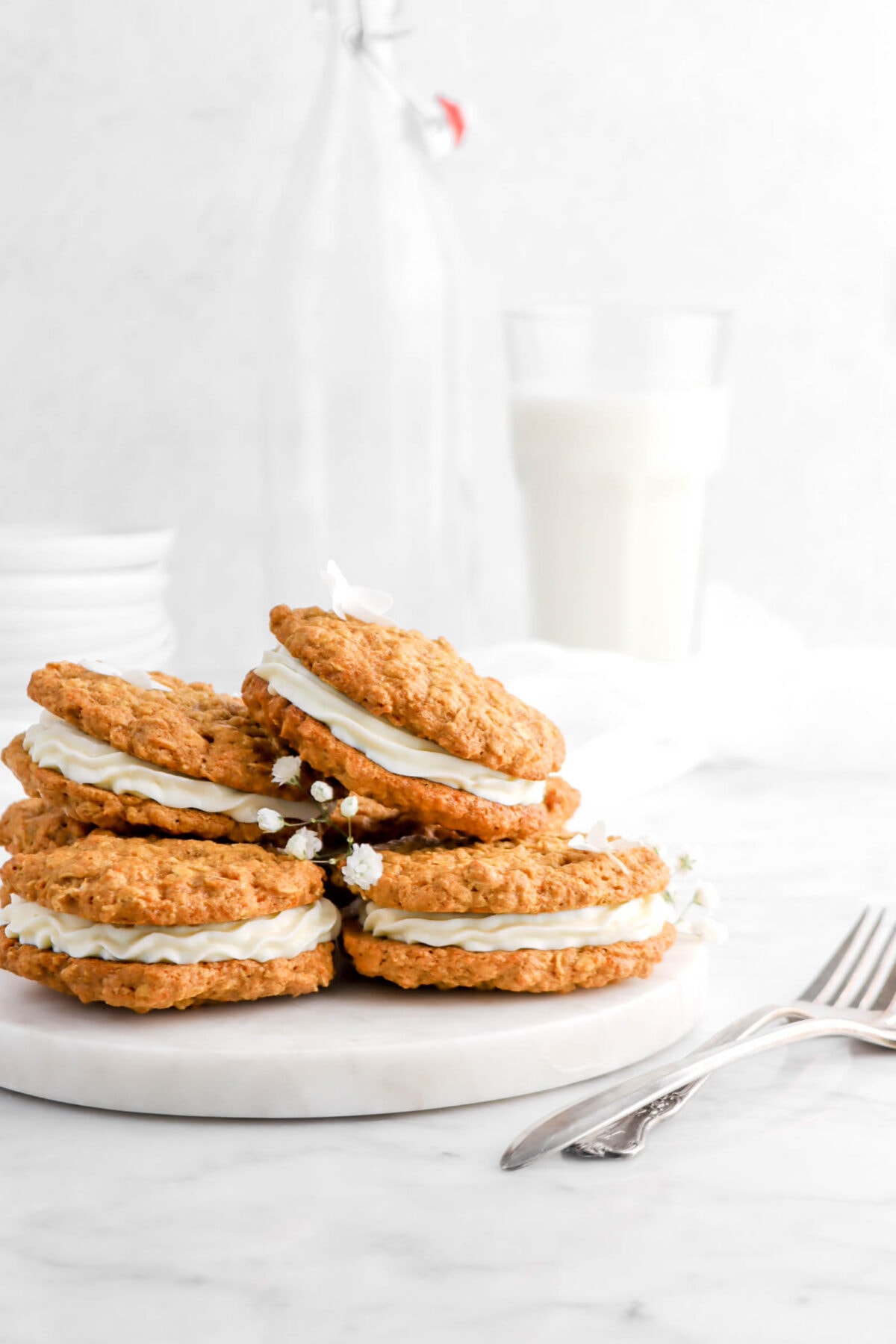 stacked carrot cake sandwich cookies on marble surface with two forks beside and glass of milk behind