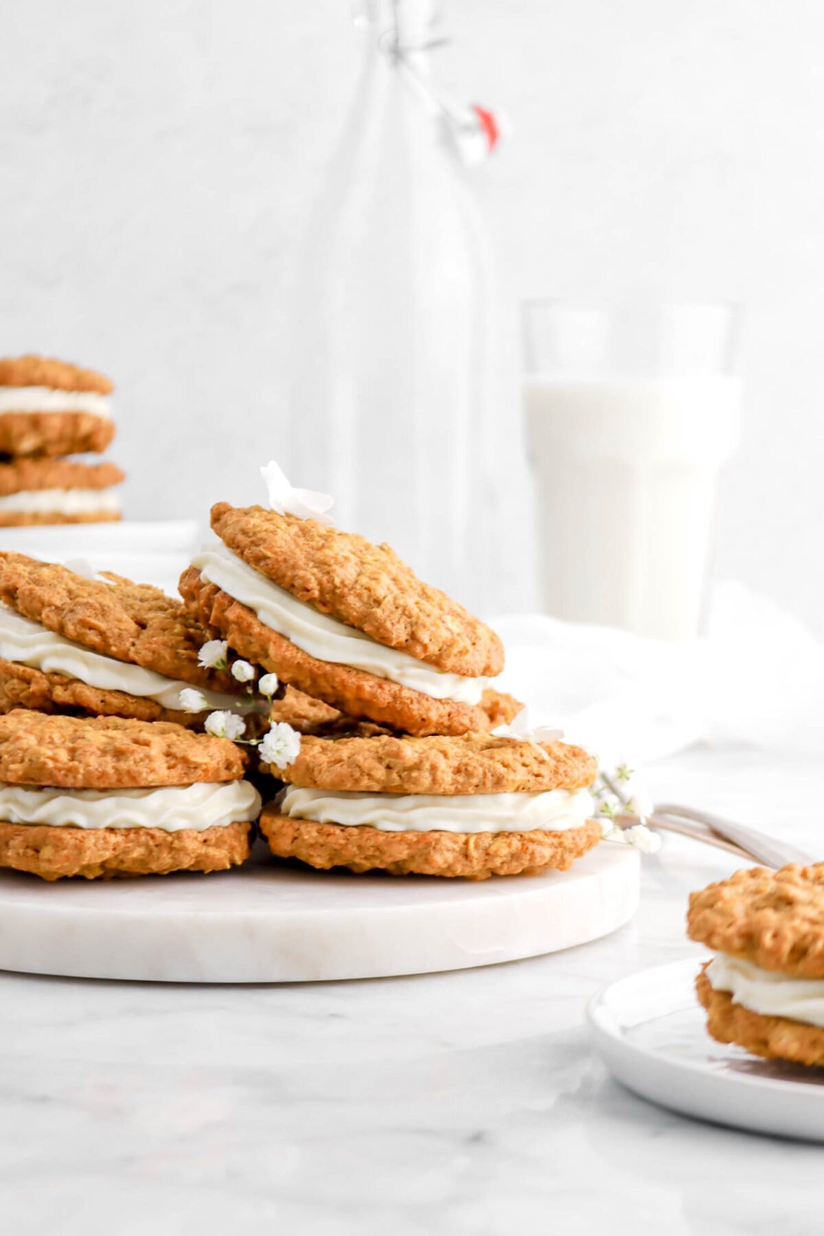 close front shot of stacked carrot cake sandwich cookies with sandwich cookies in front and behind with white flowers and glass of milk