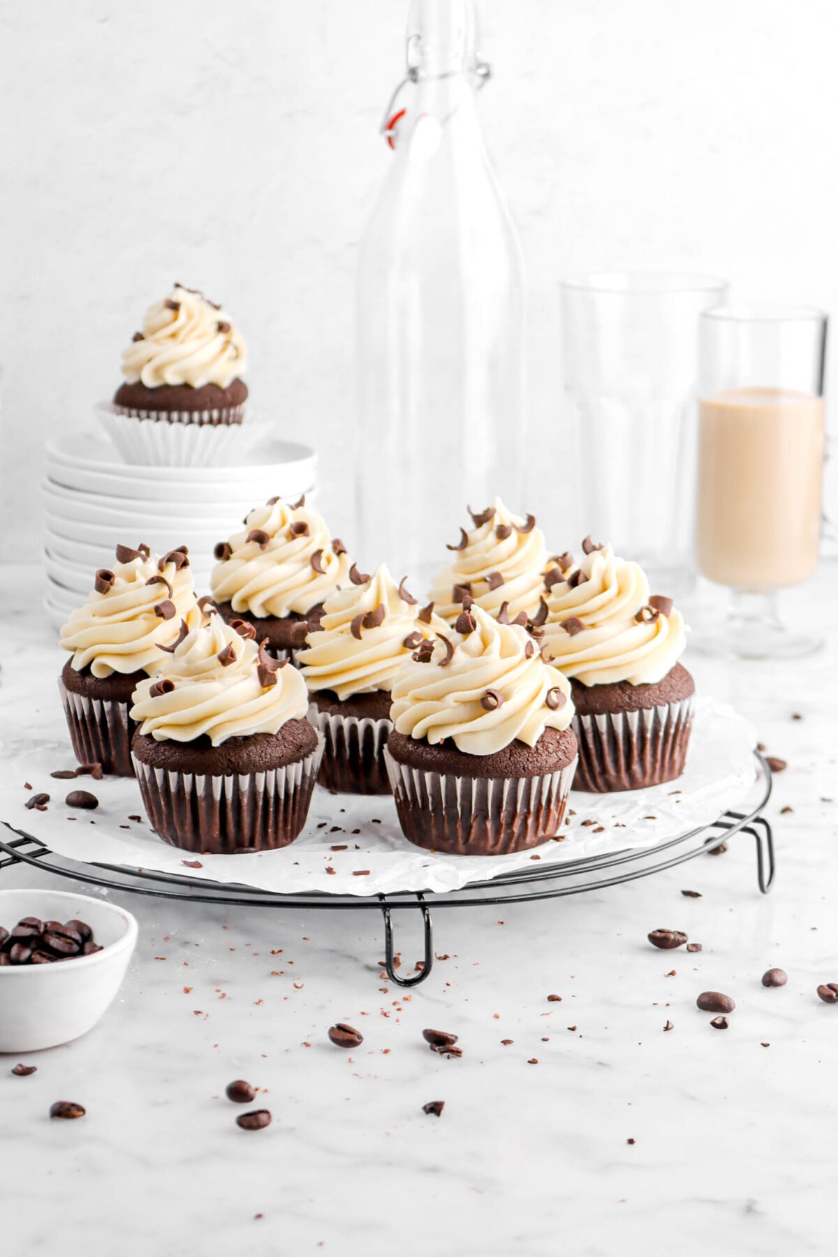 pulled back photo of five chocolate espresso cupcakes on wire cooling rack with coffee beans and chocolate curls around, a stack of white plates, two empty glasses, and a glass of irish cream behind