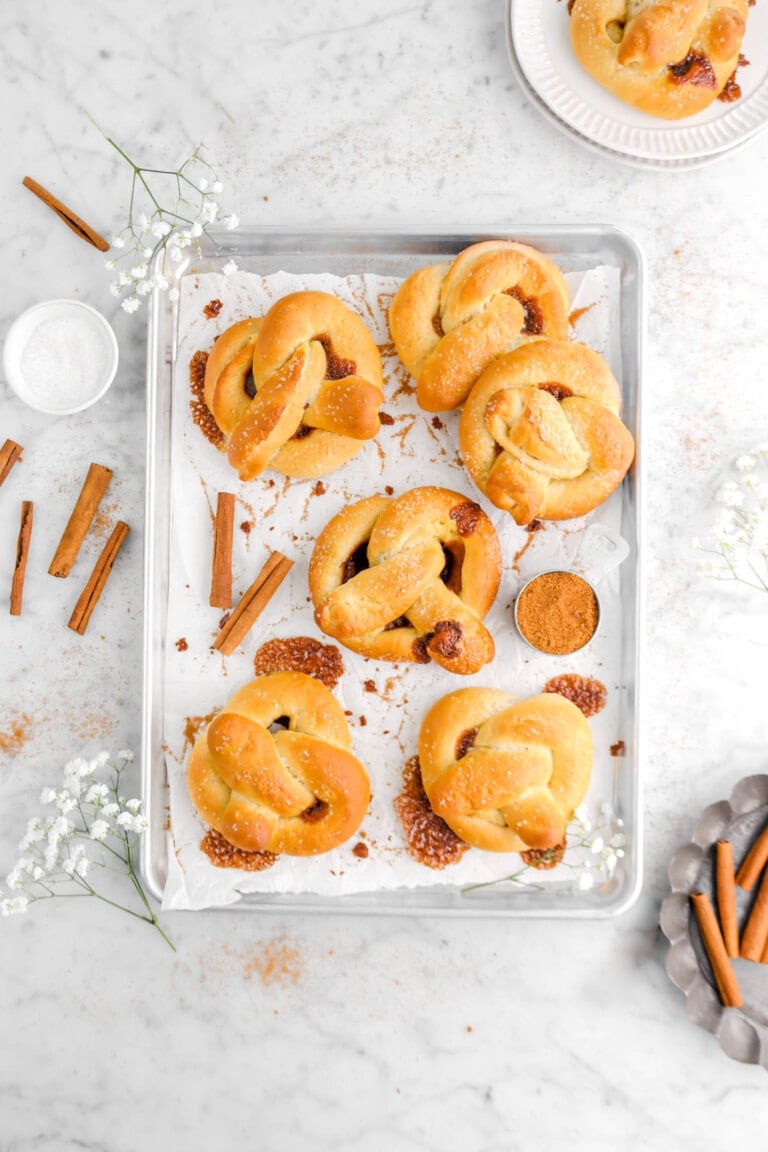 six cinnamon sugar pretzels on parchment lined sheet pan with white flowers, cinnamon sticks, and cinnamon sugar around, stack of plates with pretzel on top and old pie plate beside