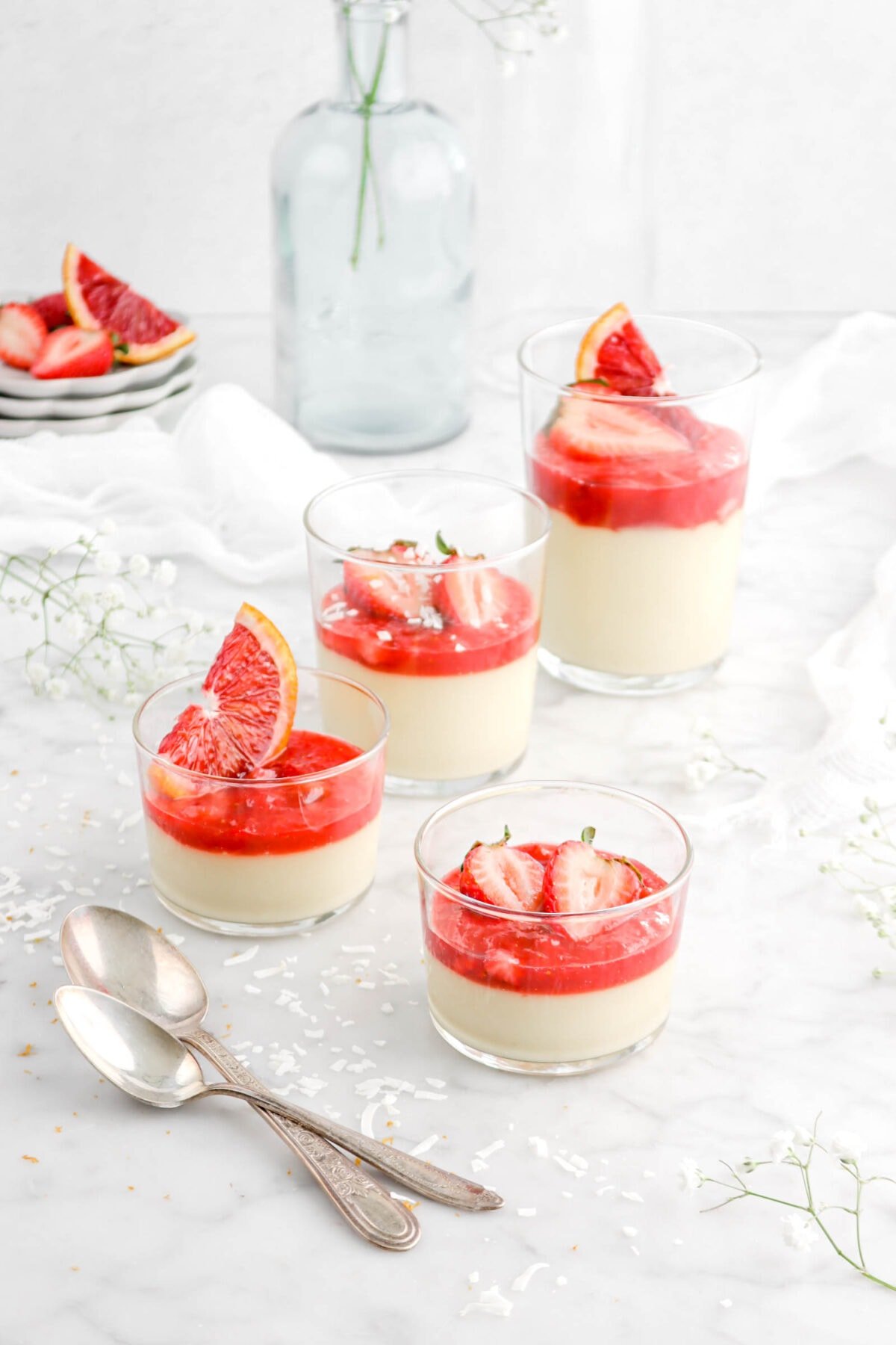 angled photo of four glasses of coconut custard with compote, sliced strawberries, and blood orange slices on top with coconut flakes and two spoons beside