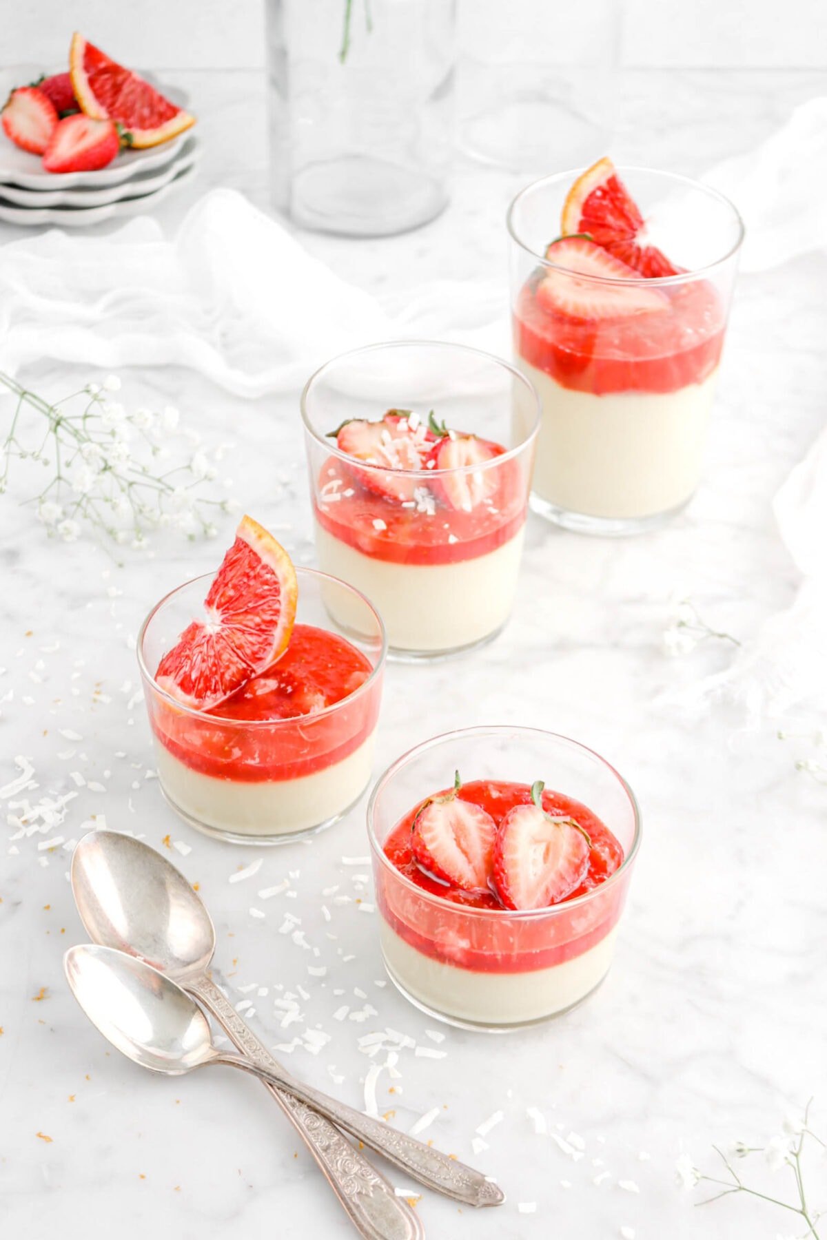 angled shot of coconut custard in four glasses with cut fruit on top, compote, and two spoons beside