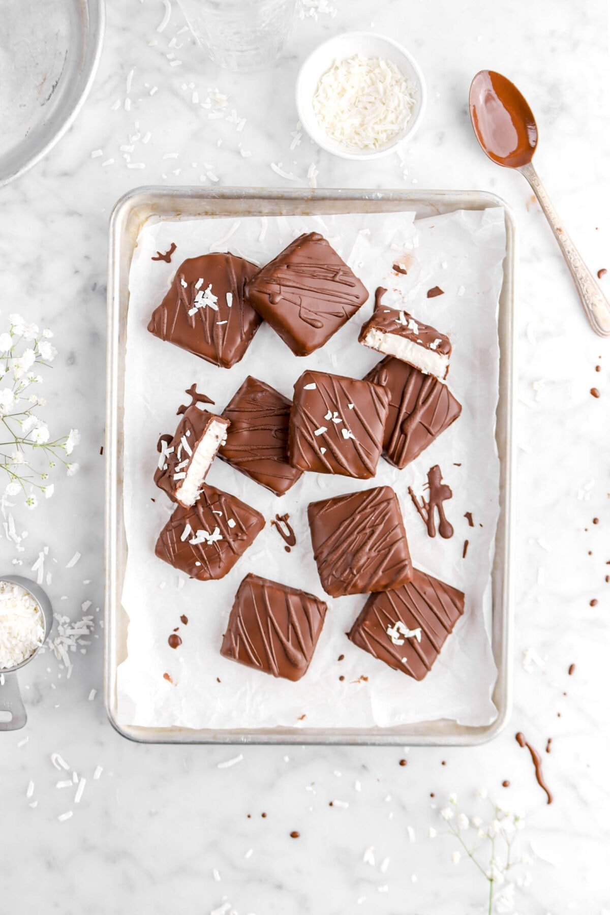 overhead shot of ten coconut filled chocolates with one cut in half, bowls of flaked coconut around, and spoonful of melted chocolate on marble surface