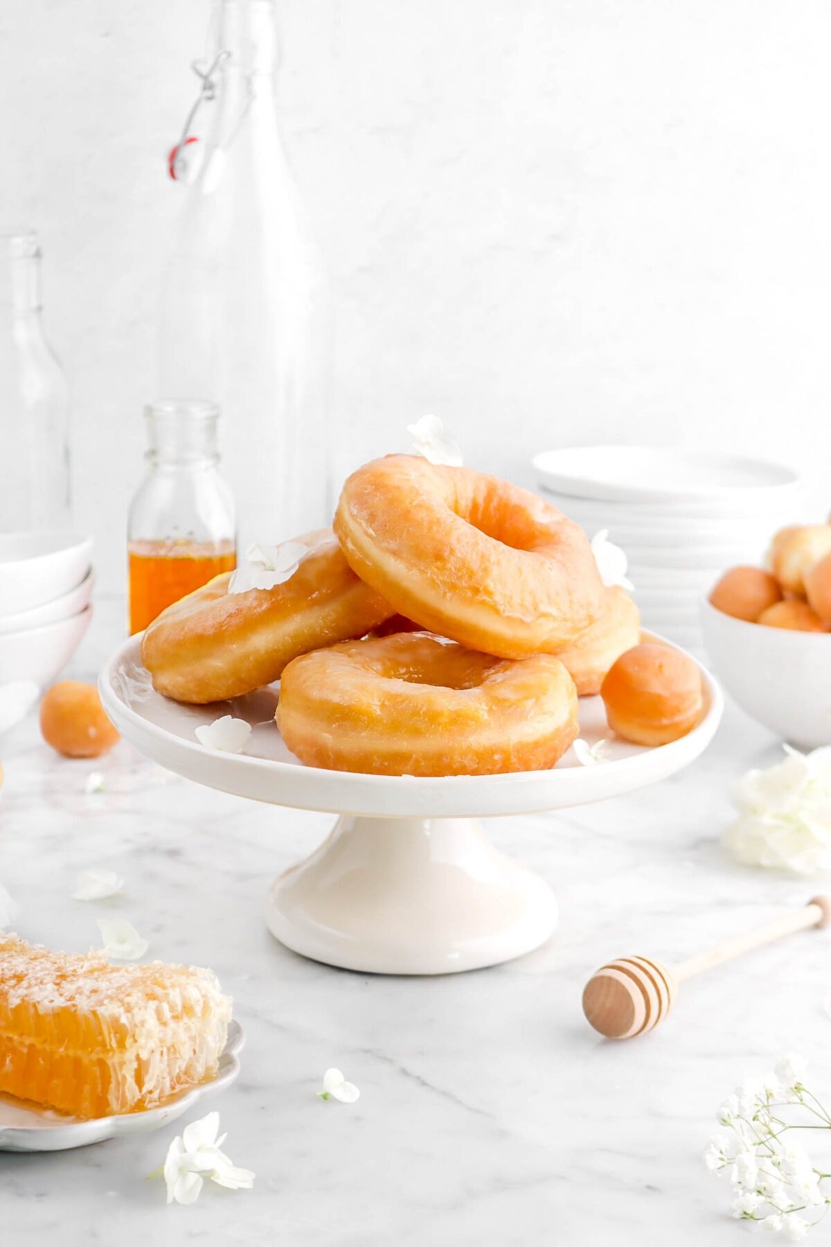 stack of honey doughnuts on cake plate with honeycomb and wooden honey dipper beside, white flowers and stack of plates behind