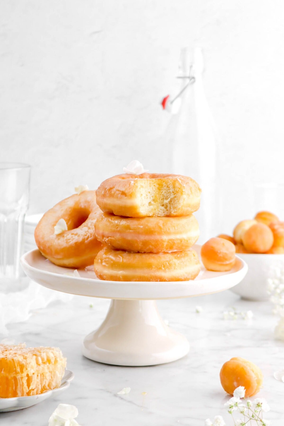 stack of three doughnuts on cake palte with bite missing out of top doughnut, another doughnut behind, and doughnut holes scattered around with white flowers
