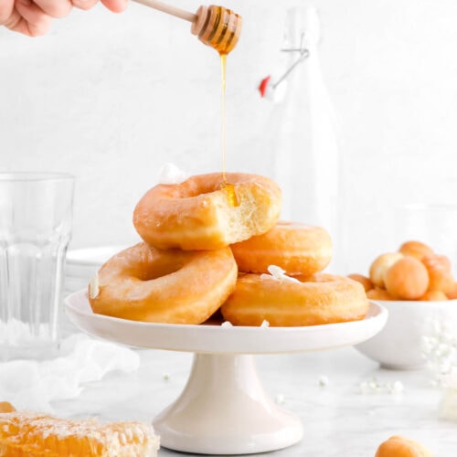honey being drizzled onto stack of doughnuts with bite missing from top doughnut, flowers, and doughnut holes around and honey comb