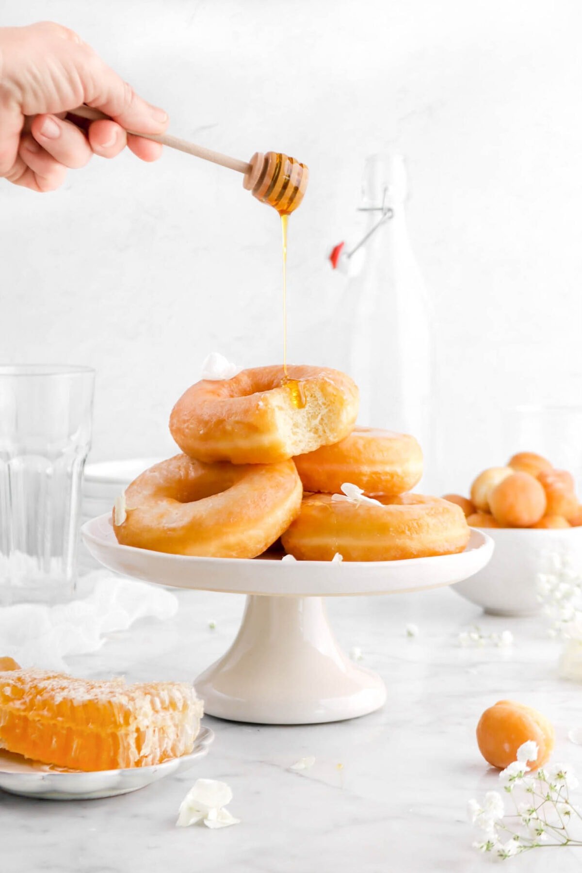 honey being drizzled onto stack of doughnuts with bite missing from top doughnut, flowers, and doughnut holes around and honey comb