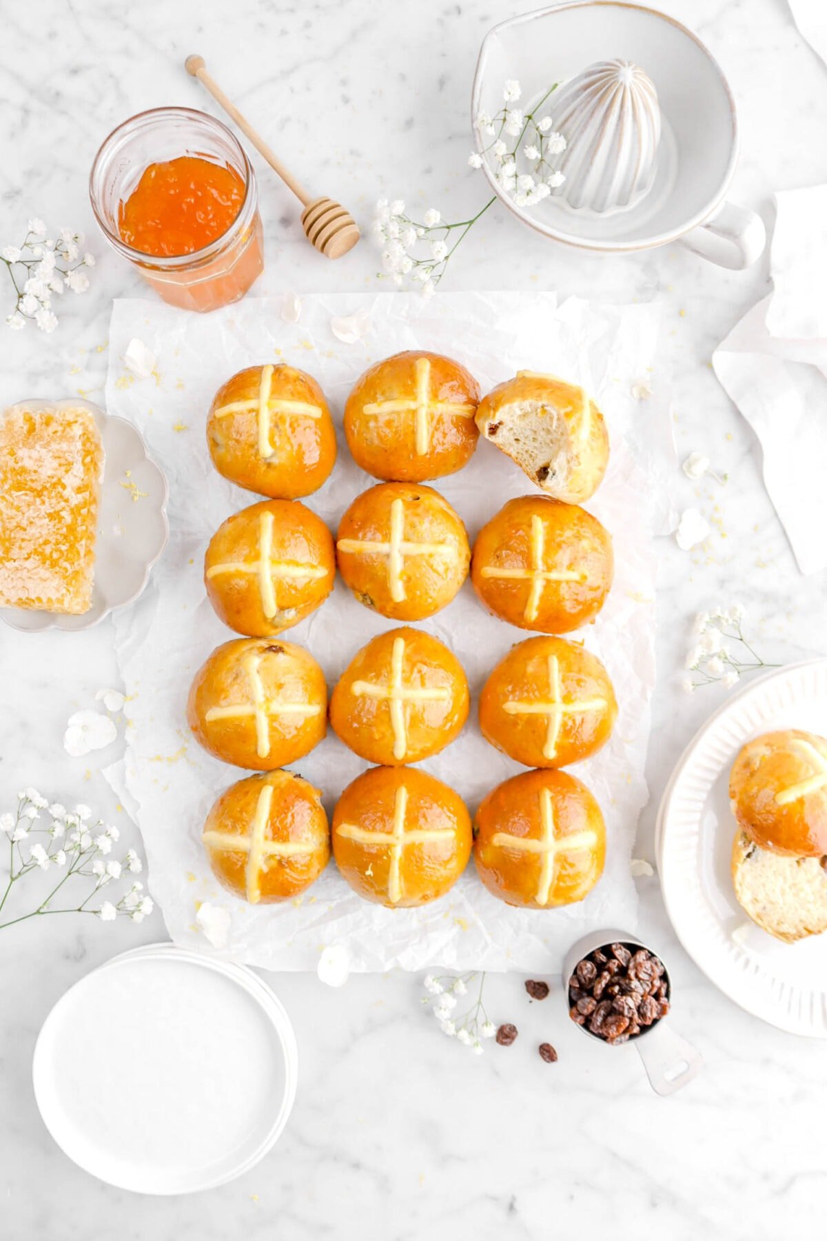 overhead of twelve hot cross buns on parchment paper with bite taken out of top right bun with white flowers around, a homey comb, jar of jam, citrus juicer, and measuring cup full of raisins
