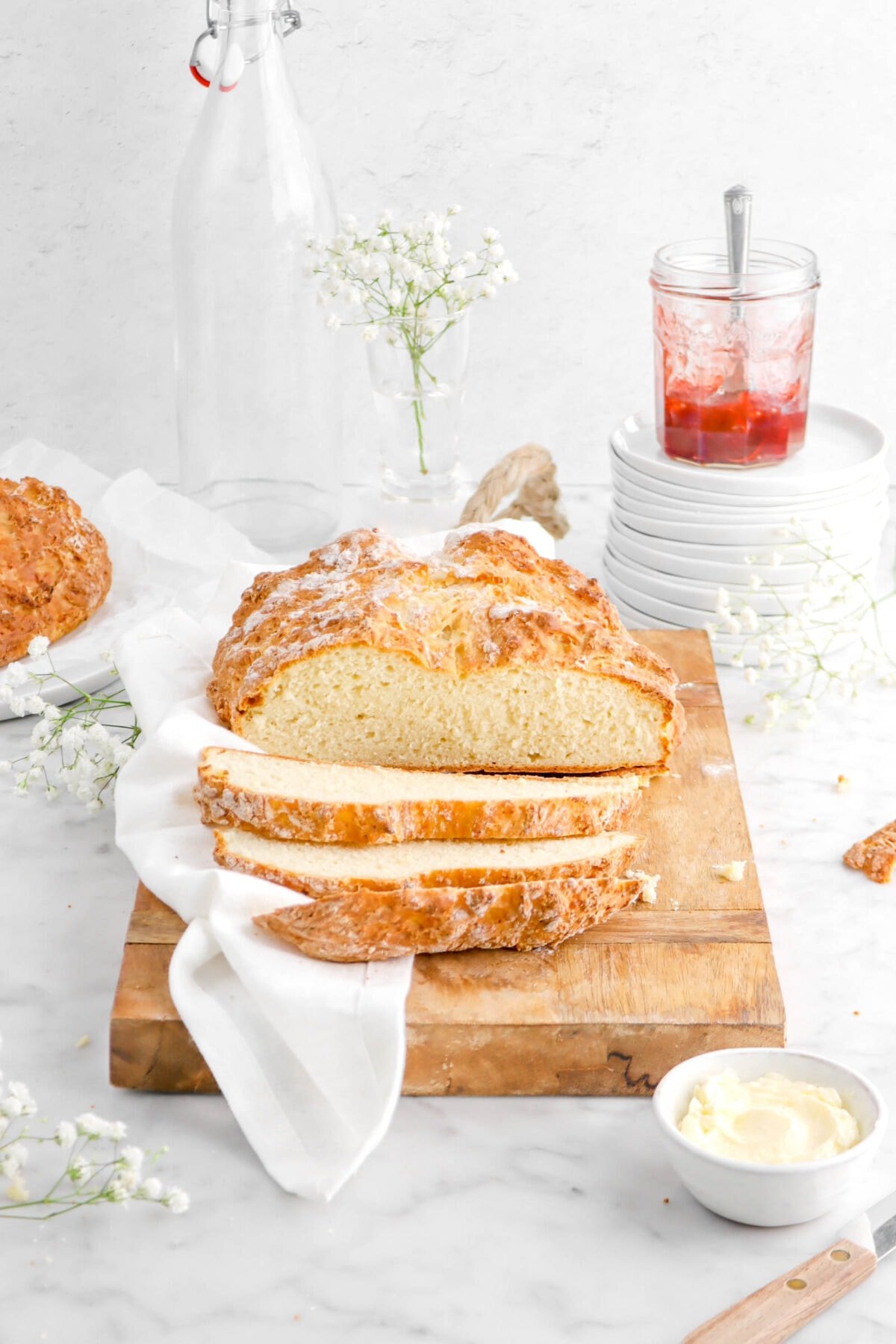 angled photo of sliced irish soda bread on wood board with white napkin, flowers, bowl of butter, crumbs, stack of white plates, and jar of jam behind