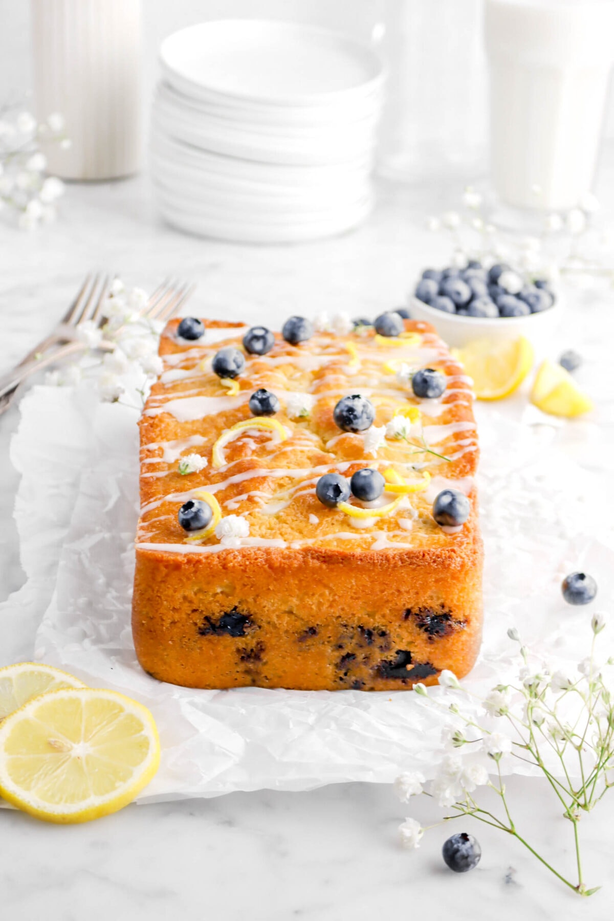 front photo of loaf cake on parchment paper with blueberries, lemon slices, stack of plates, and glass of milk behind with flowers around