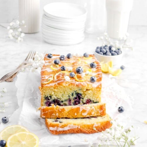 lemon loaf cake on parchment paper with two slices laying in front, flowers, lemon slices, and blueberries around with stack of plates and glass of milk behind