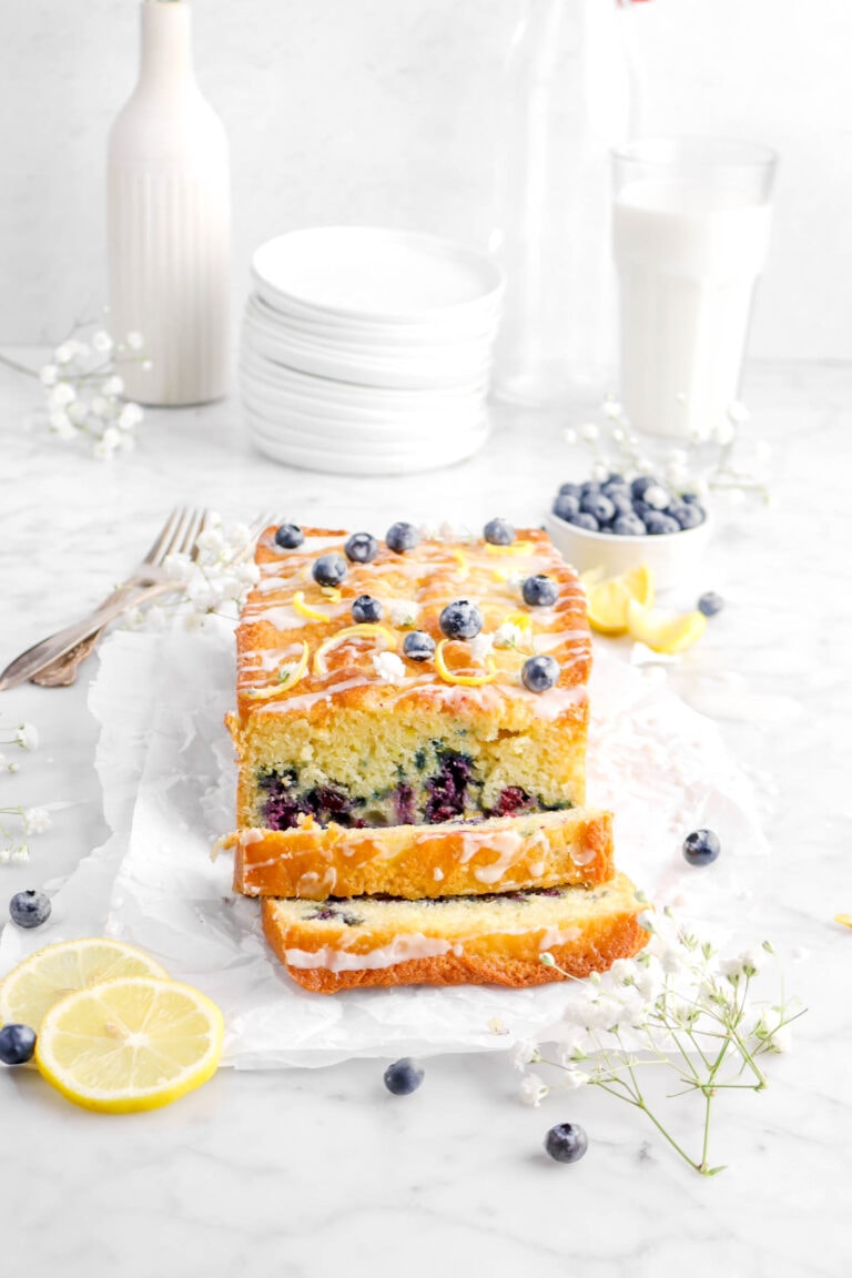 lemon loaf cake on parchment paper with two slices laying in front, flowers, lemon slices, and blueberries around with stack of plates and glass of milk behind