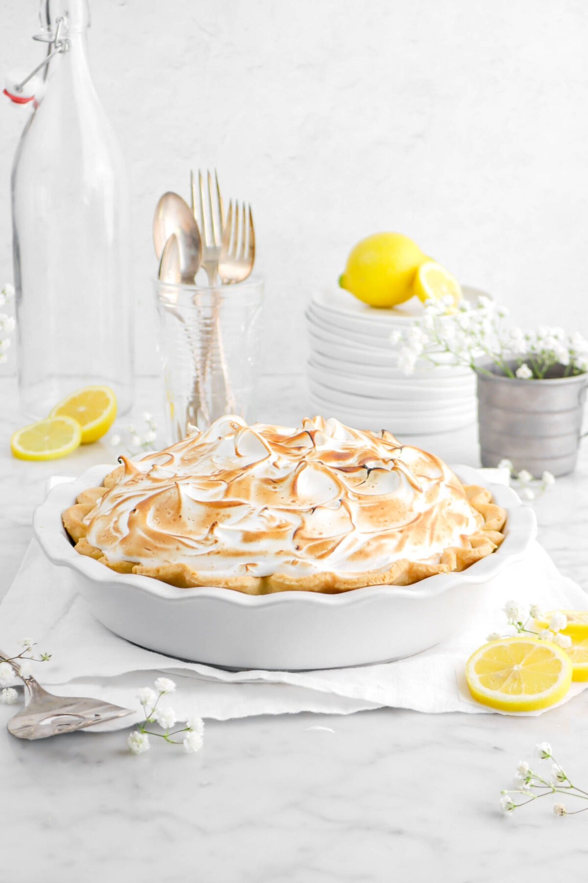 front shot of lemon meringue pie on white napkin with lemon slices, flowers, stack of plates, and utensils standing up in a glass