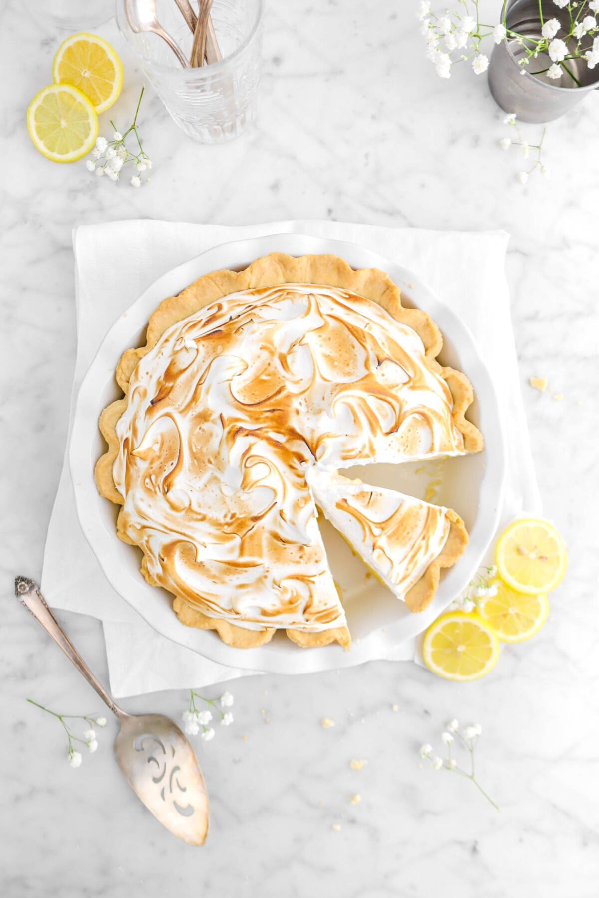 overhead shot of lemon meringue pie on white napkin with cake knife with white flowers
