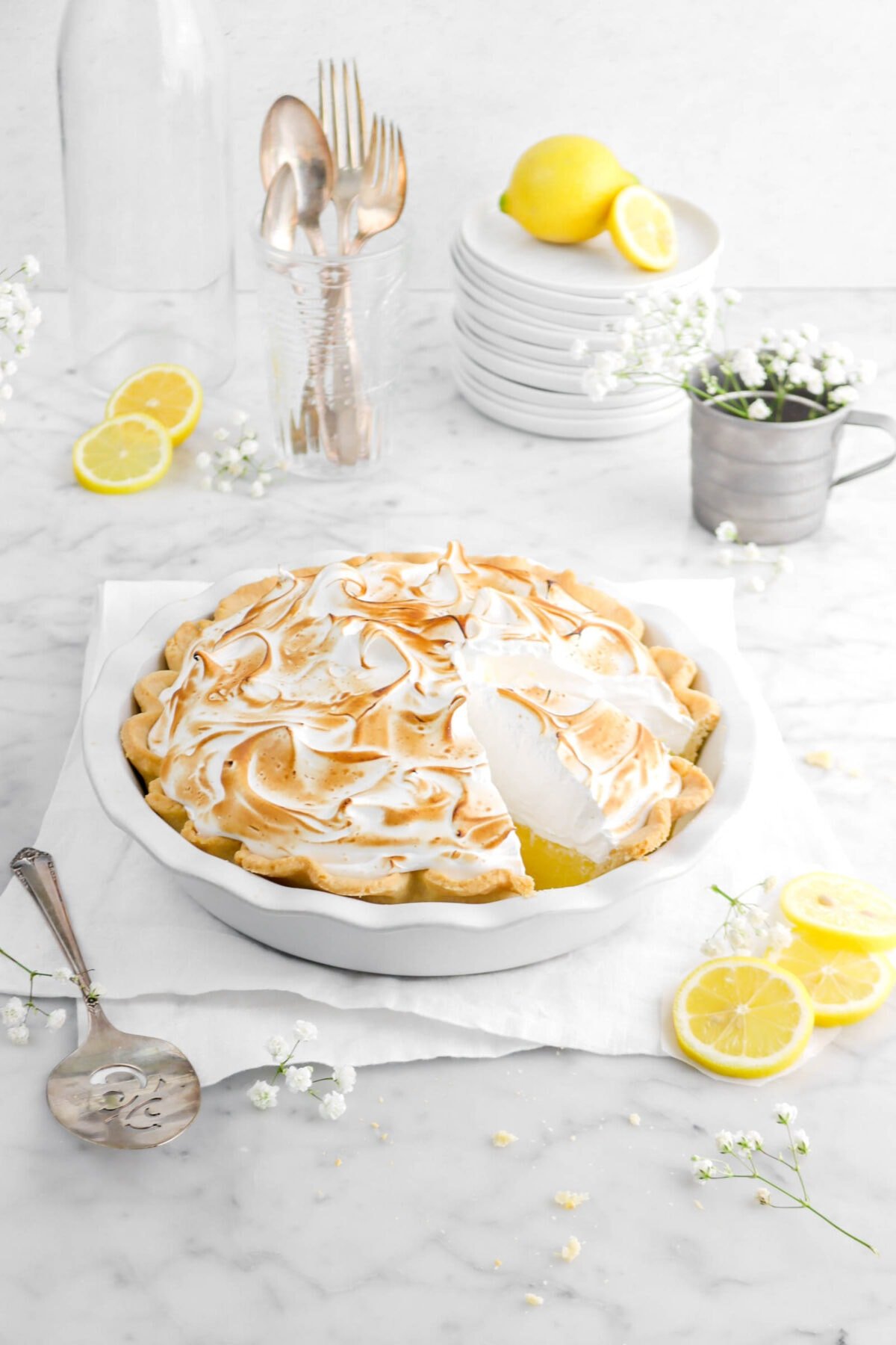 pulled back shot of meringue pie with flowers, lemons, and stack of white plates behind