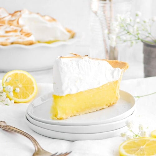 meringue pie slice on stack of white plates with fork beside on white napkin with lemon slices and meringue pie behind