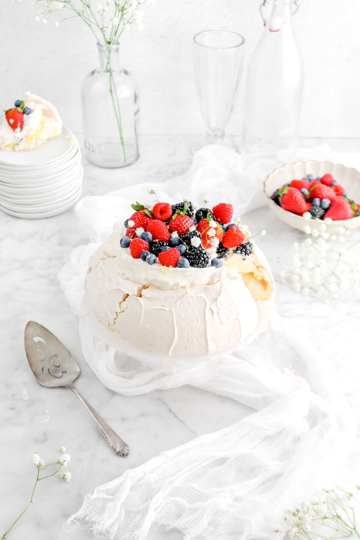angled photo of pavlova with slice on stack of white plates behind, mixed berries in a bowl, white cheese cloth underneath cake plate, and white flowers around