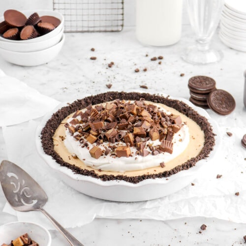 angled photo of peanut butter pie with whipped cream and chopped peanut butter cups on top with glass of milk and stack of plates behind and bowl of peanut butter cups