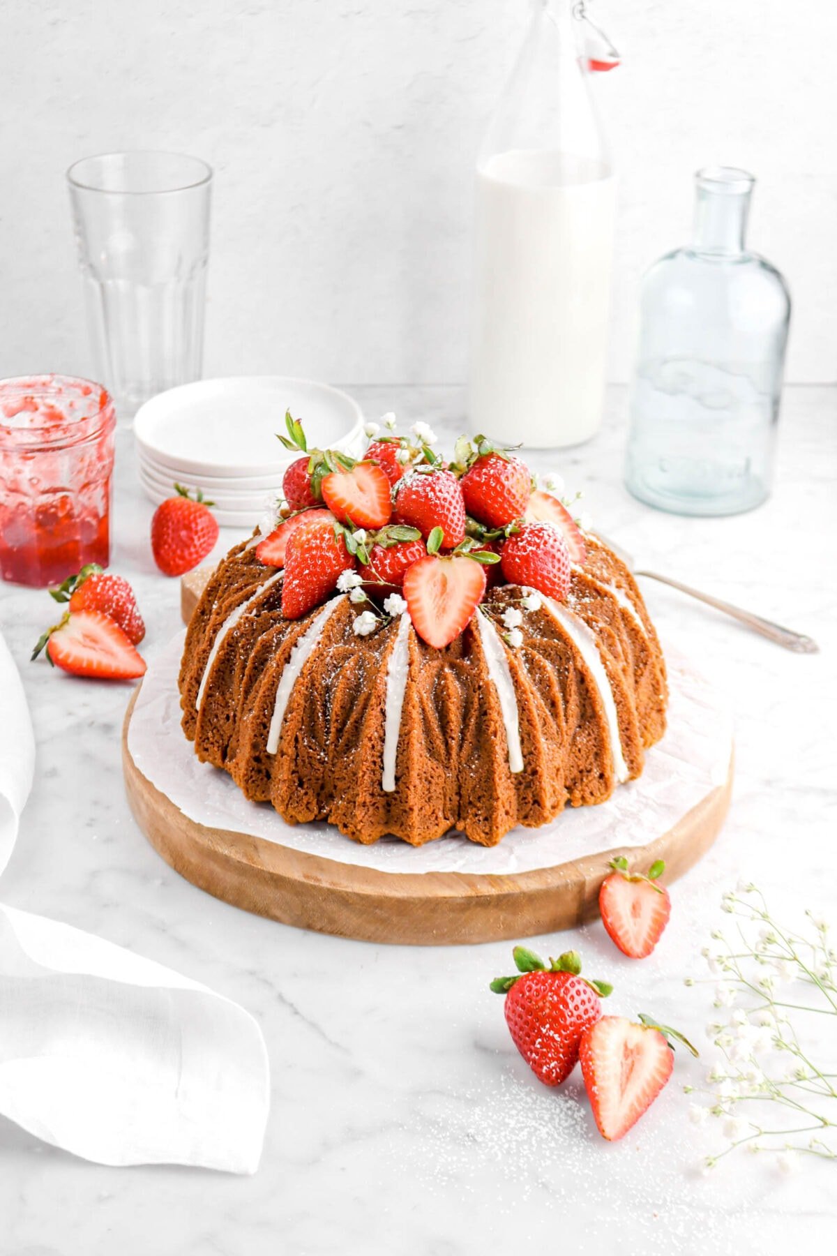 angled photo of bundt cake on wood board with icing dripping, fresh strawberries, white flowers, and powdered sugar on top, jam jar, stack of plates, empty vase, and jug of milk behind