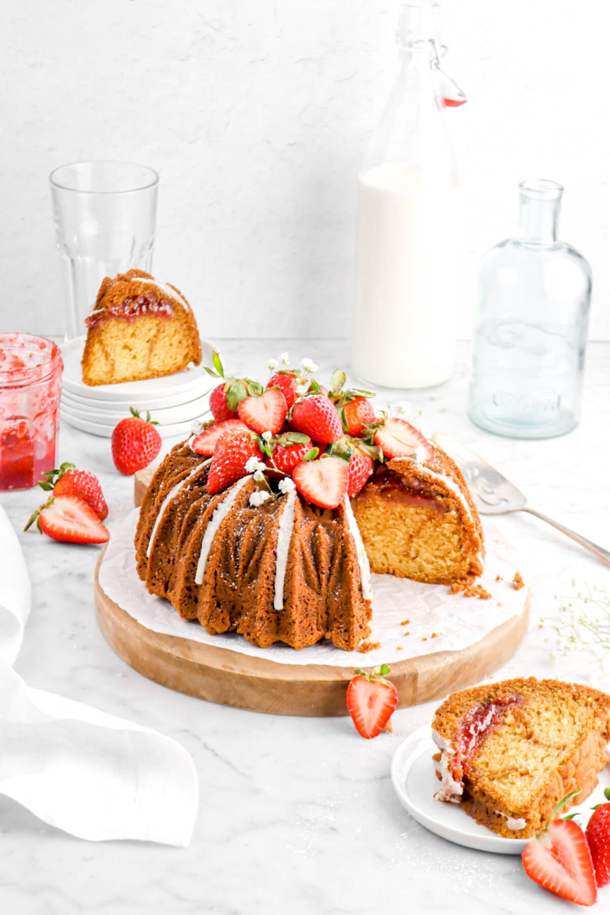 front shot of strawberry swirl bundt cake with slice in front and behind on white plates with empty vase, jug of milk, empty glass, and jam jar behind