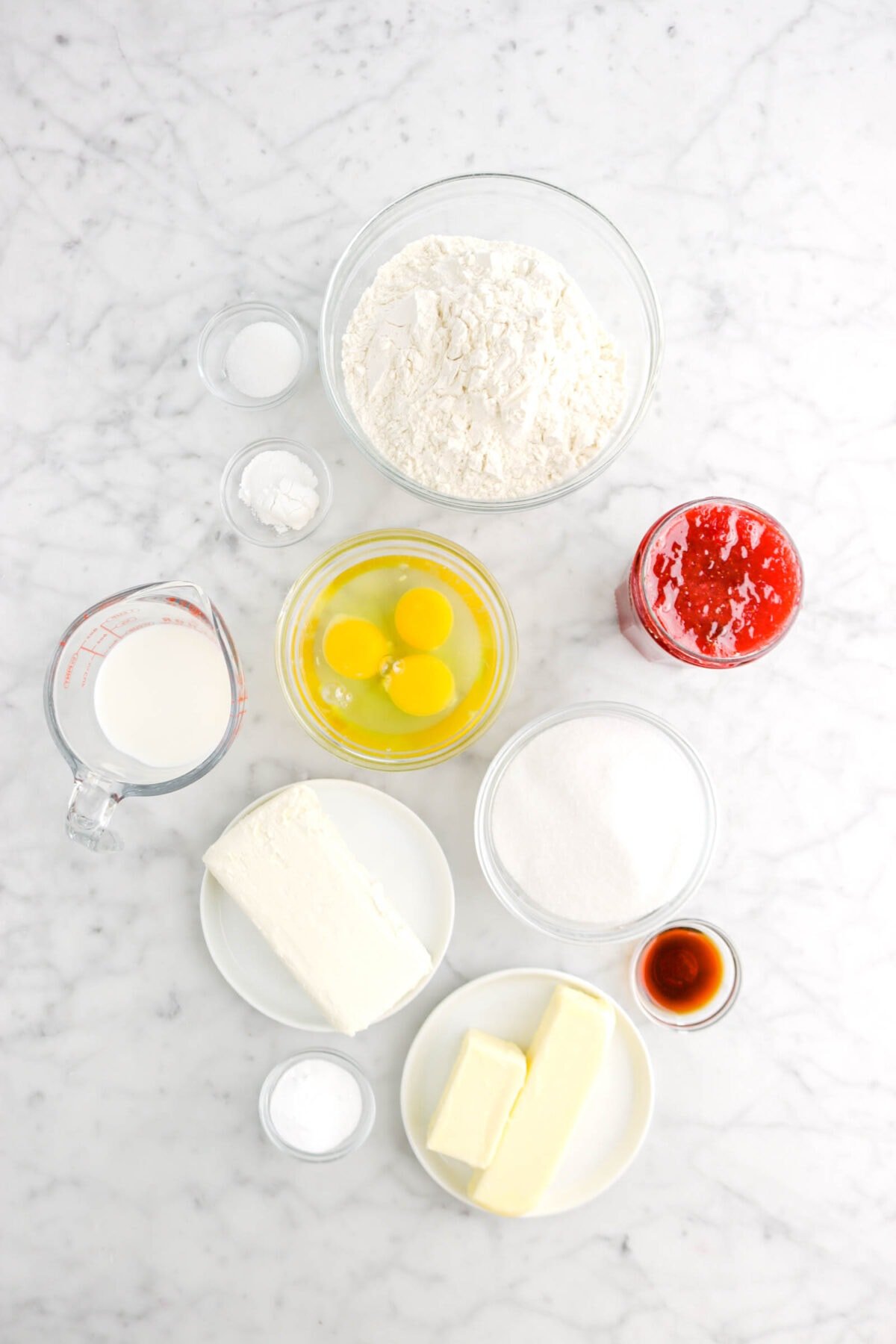 flour, baking powder, baking soda, eggs, milk cream cheese, sugar, strawberry jam, vanilla, butter, cream cheese, and salt on marble surface