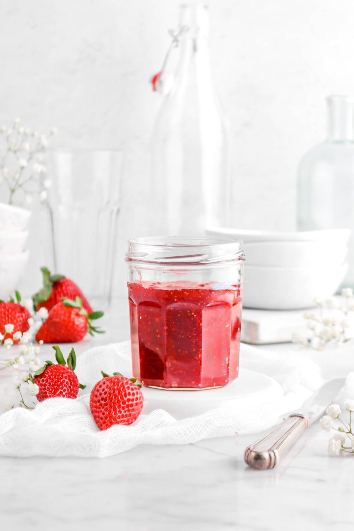 strawberry jam in glass jar on upside down white plate with white cheese cloth, silver knife, fresh strawberries, flowers, and empty glasses behind