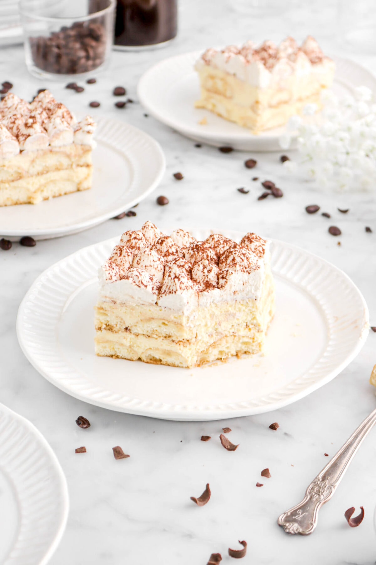 angled photo of slice of tiramisu on white plate with bite missing on marble surface with chocolate curls around and two slices of tiramisu behind