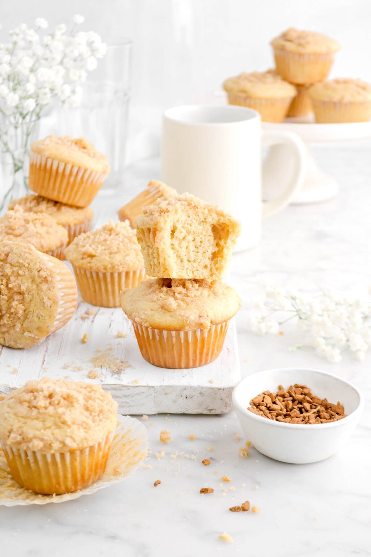 chai tea muffins on white wood board with two in the front stacked, a bit missing from one, with bowl of cinnamon chips, a coffee mug, a cake plate, and flowers behind