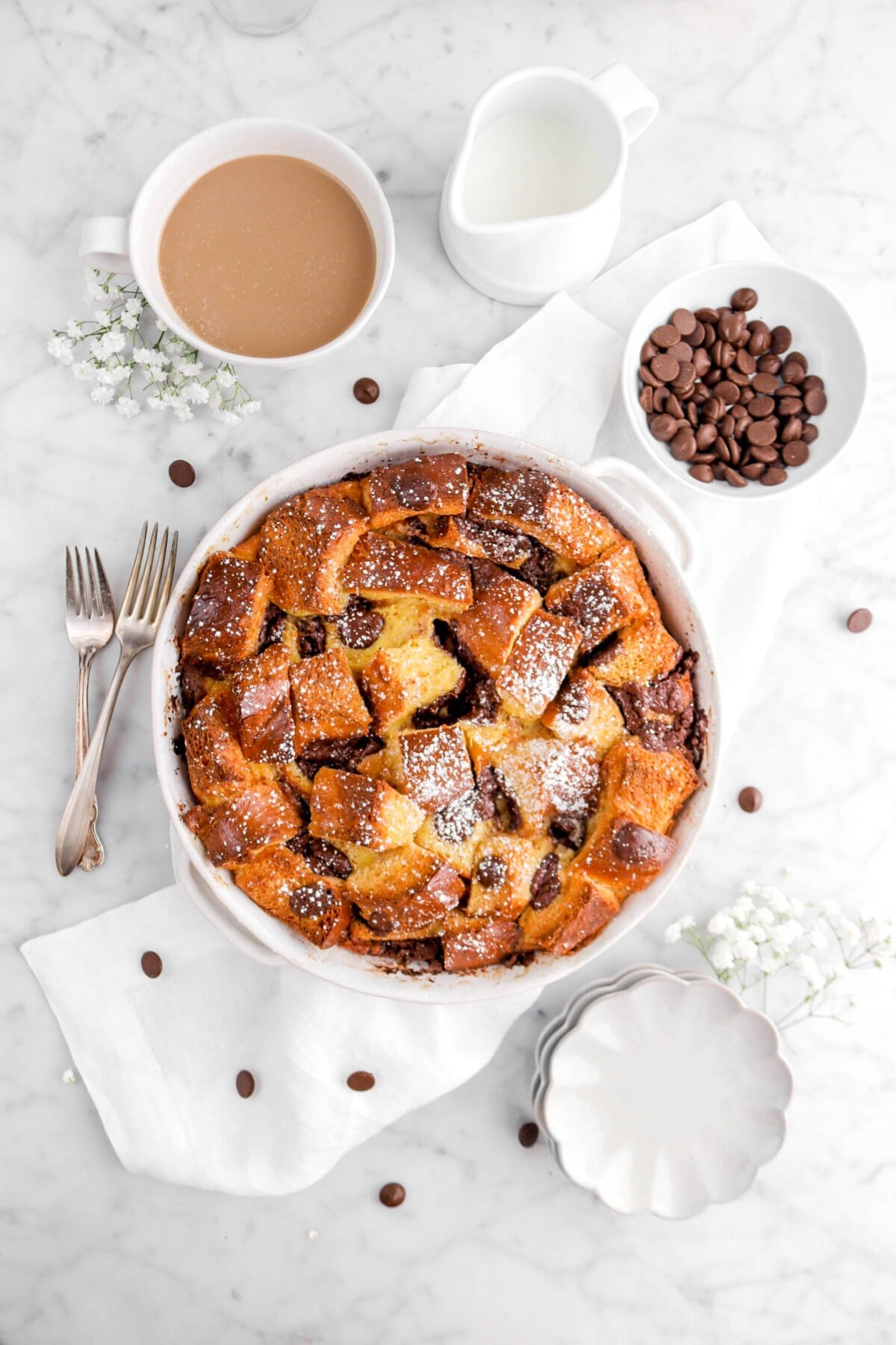 overhead shot of chocolate chip bread pudding in round casserole on white napkin with two forks, flowers, bowl of chocolate chips, cup of coffee, and white creamer beside with scalloped plates