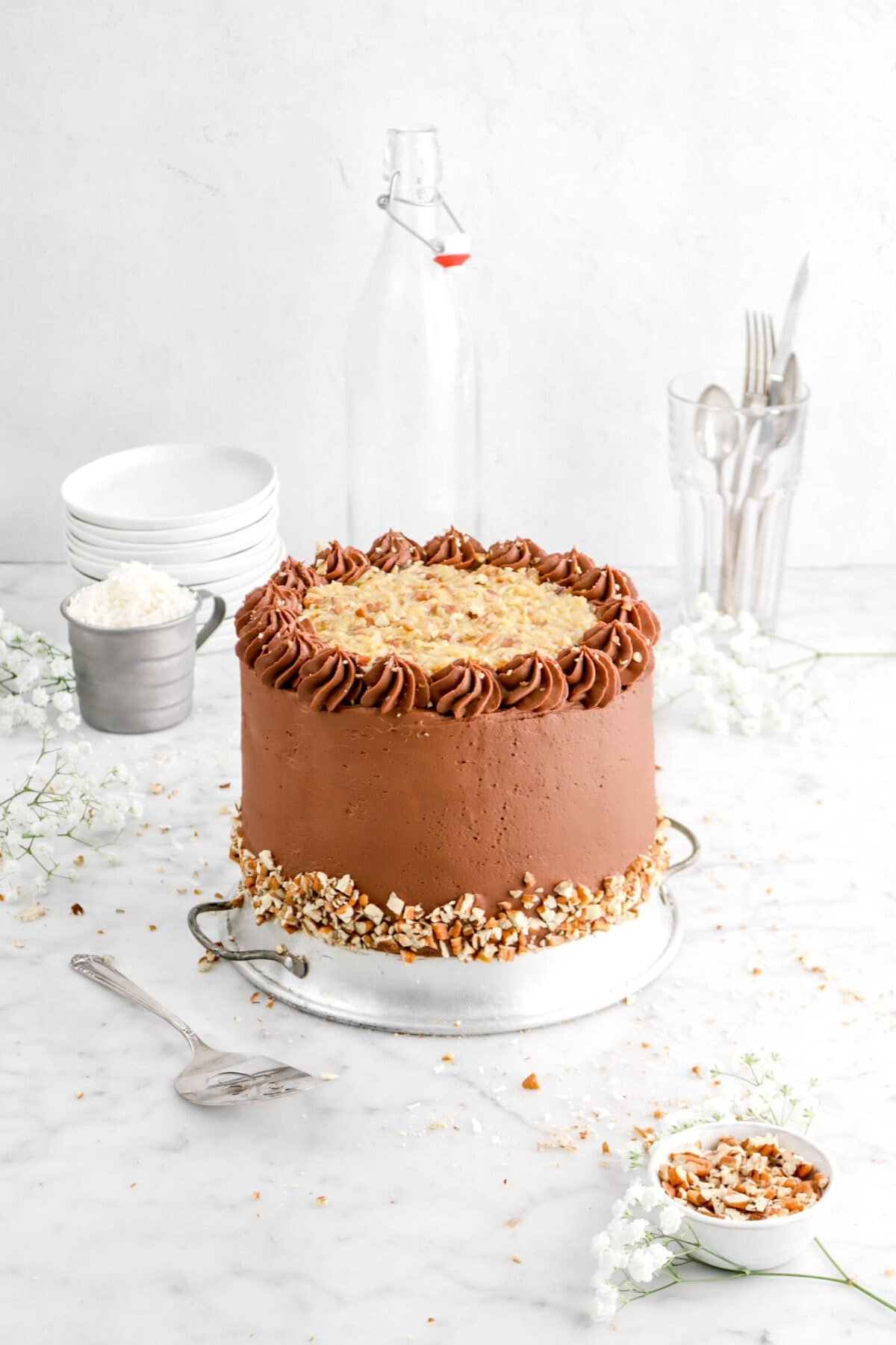 angled shot of german chocolate cake on upside down pie plate with cake knife, flowers, cup measure of coconut flakes, stack of plates, and glass full of silver utensils behind