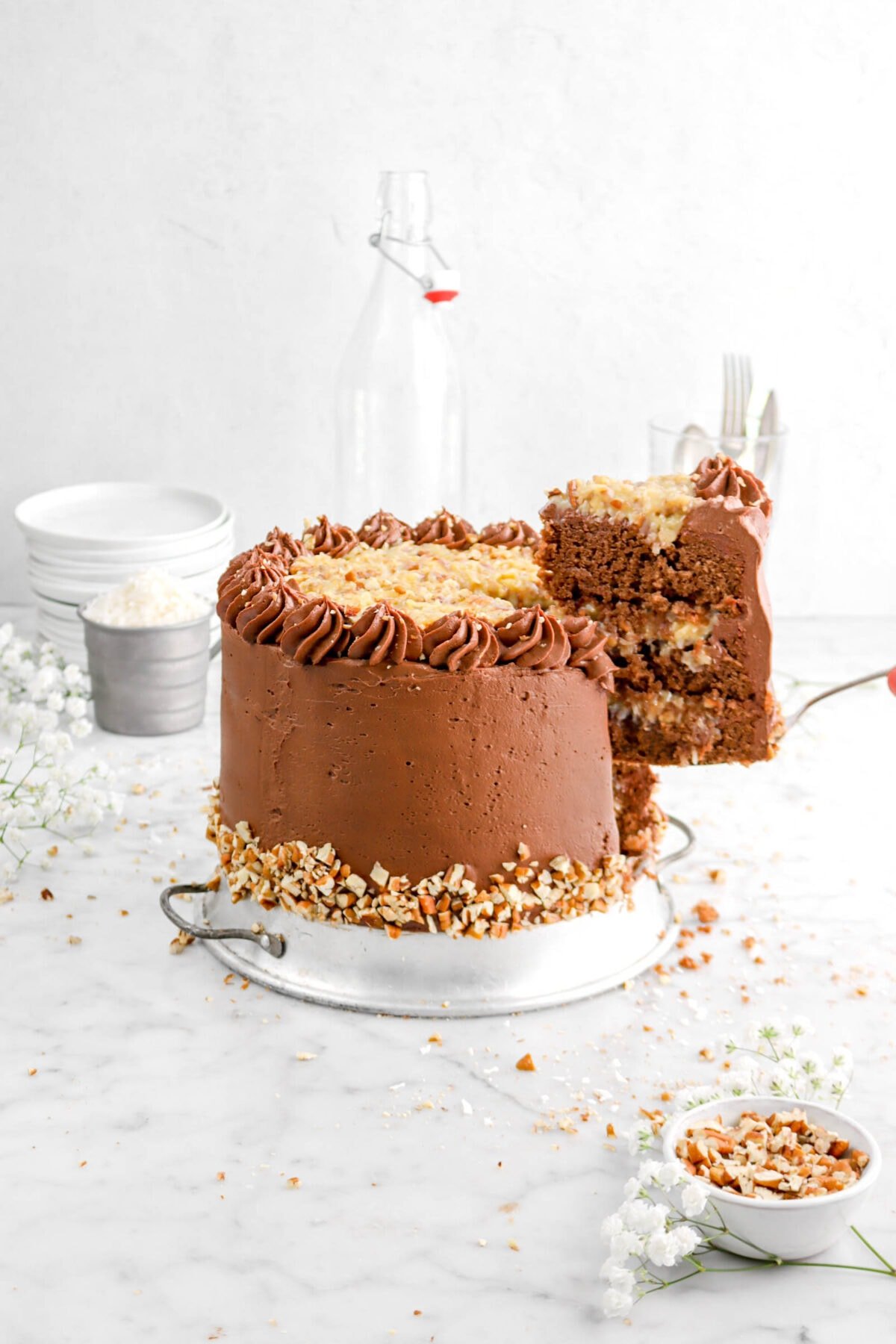 slice of german chocolate cake being held by the cake with flowers around, a bowl of chopped pecans, a cup measure of shredded coconut, and stack of white plates
