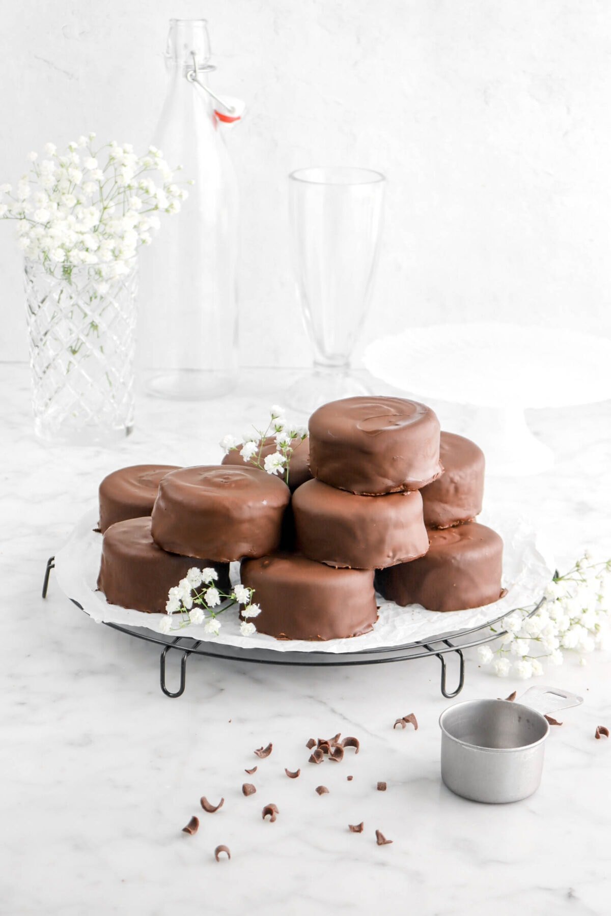 pulled back photo of mini chocolate coated cakes on wire cooling rack with cake plate behind, empty glasses, and white flowers