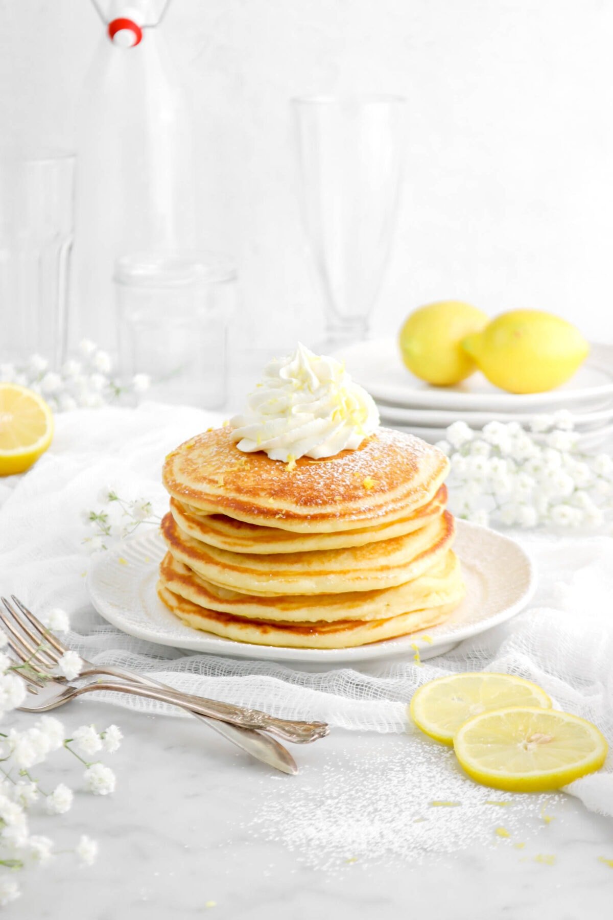 stack of pancakes on white plate with piped whipped cream on top, lemon zest, and powdered sugar with lemon slices around, two forks, and flowers