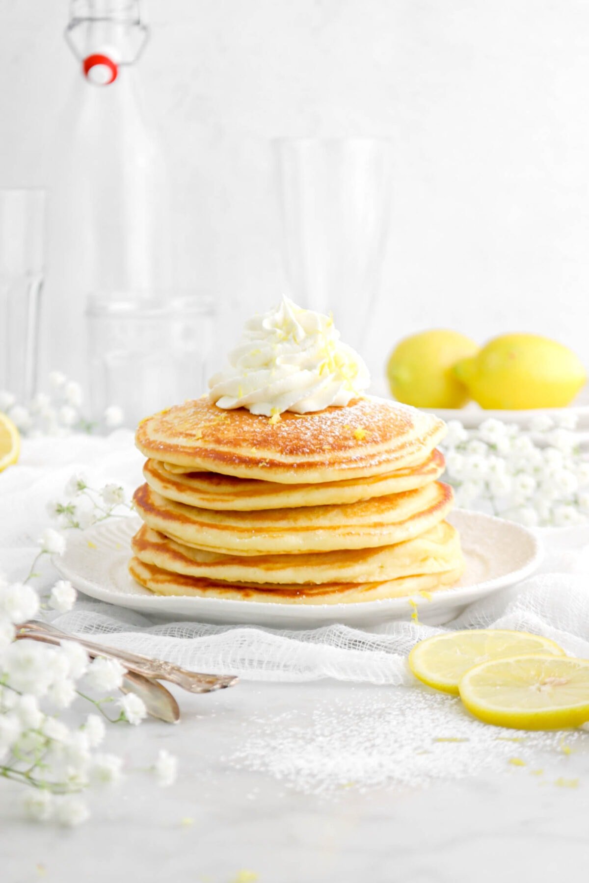 five stacked pancakes on white plate with flowers around, lemon zest, piped whipped cream, and powdered sugar on top of pancakes with two whole lemons behind