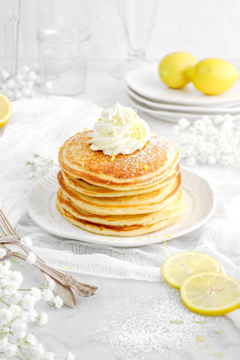 angled close up of five stacked lemon ricotta pancakes with piped whipped cream, lemon zest, and powdered sugar on top, lemon slices beside, and two forks