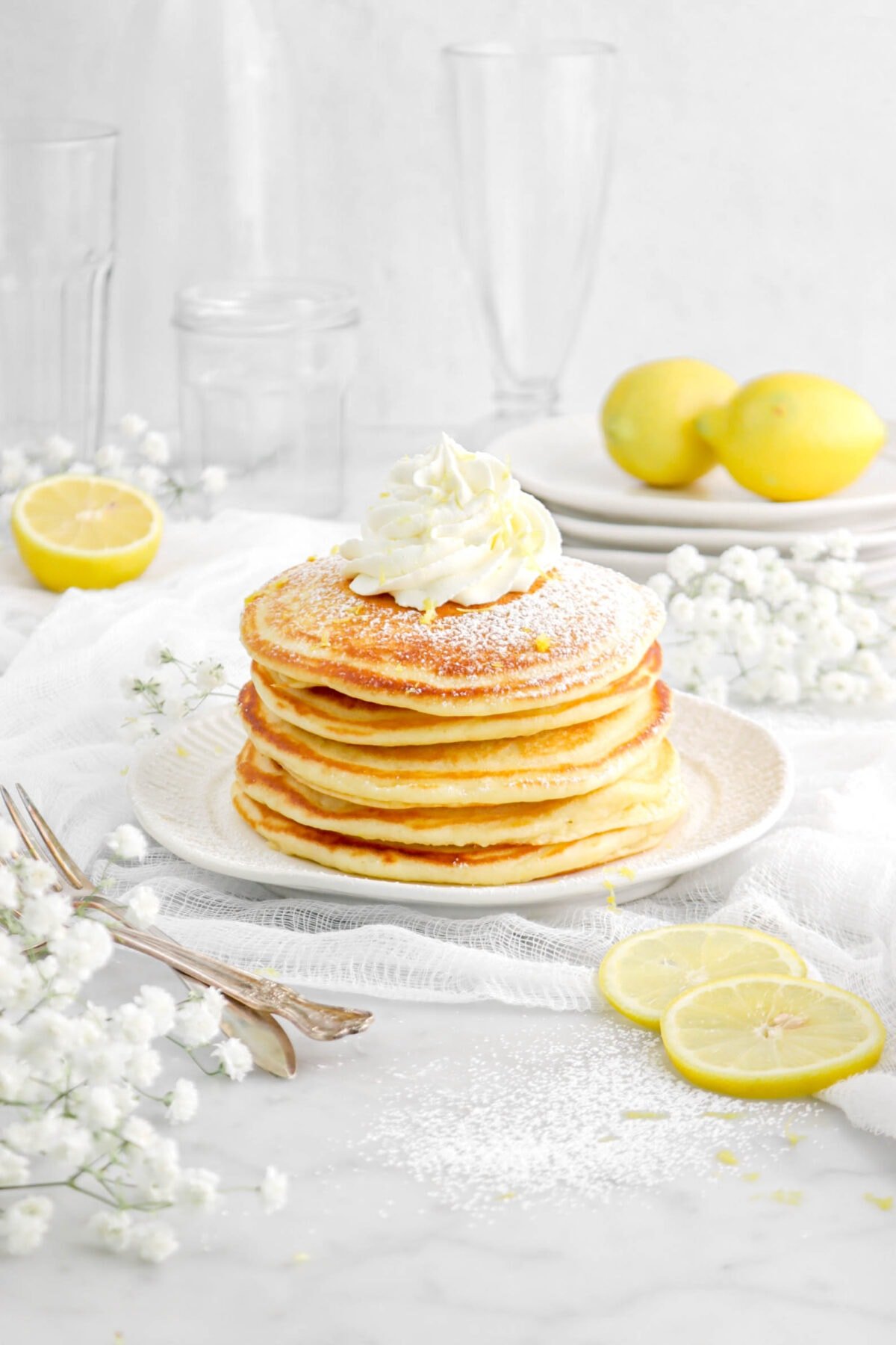 lemon ricotta pancakes stacked on white plate with whipped cream and powdered sugar on top, flowers, two forks, and lemon slices beside