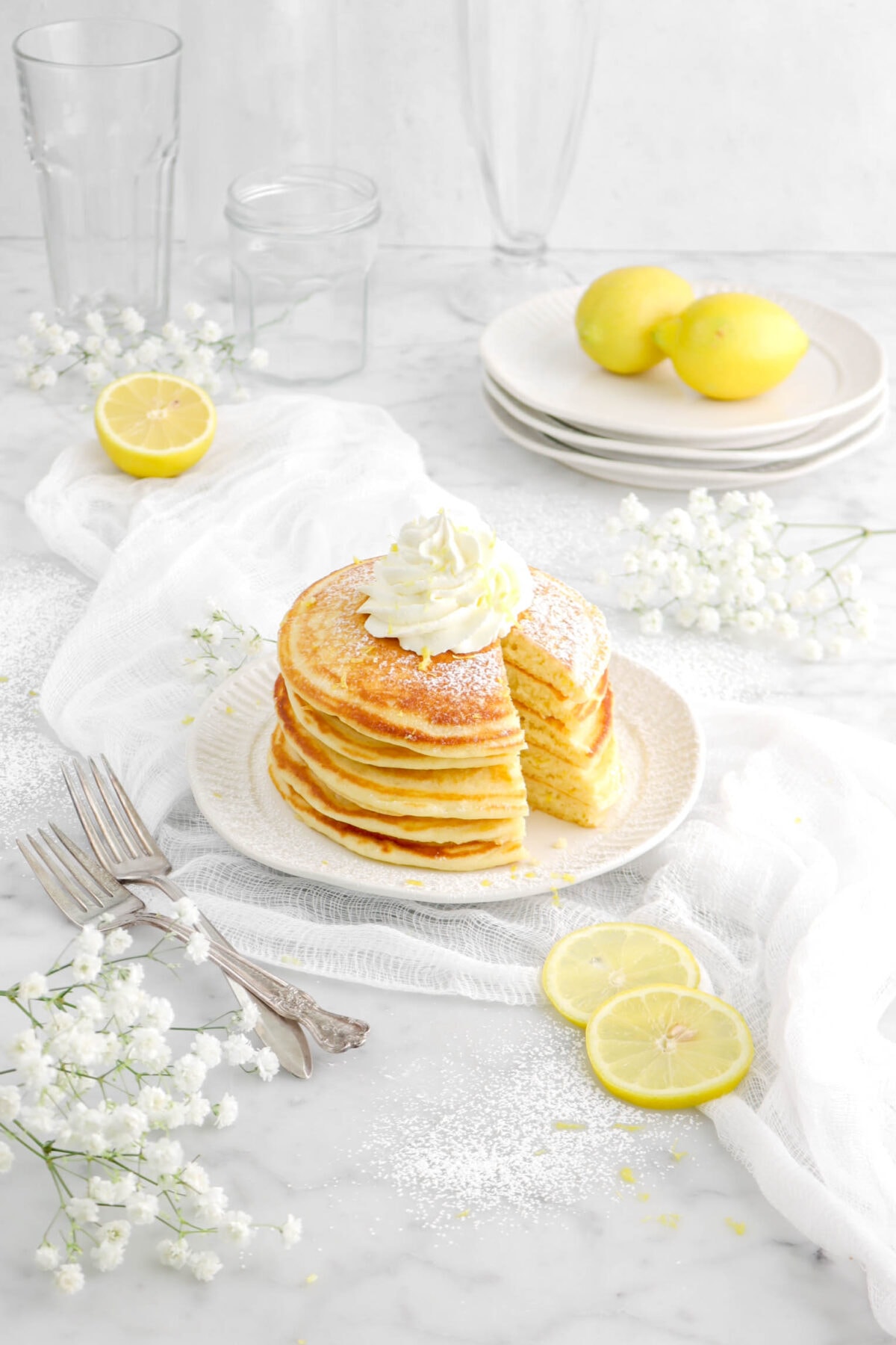 pulled back shot of sliced lemon ricotta pancakes on white plate with lemons, forks, white cheese cloth, and flowers around