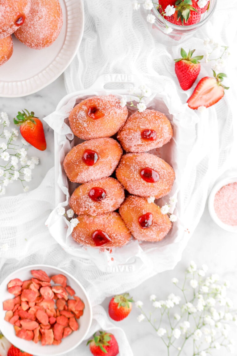 overhead shot of seven jam filled doughnuts in white casserole with plate of doughnuts beside, bowl of dried strawberries, and flowers around
