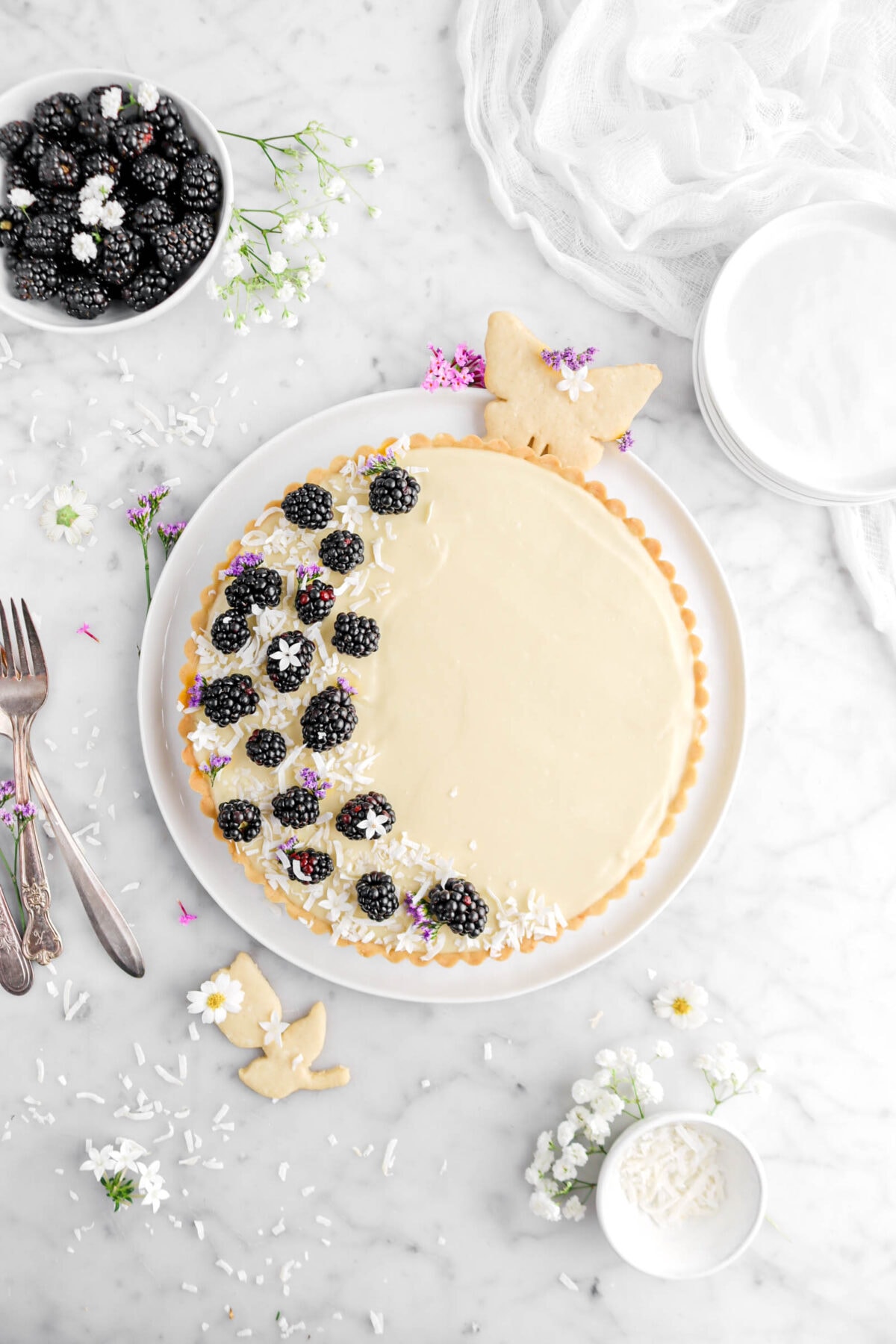 coconut blackbery tart on white plate with a butterfly and tulip cookie beside, forks, coconut flakes, and bowl of fresh blackberries beside