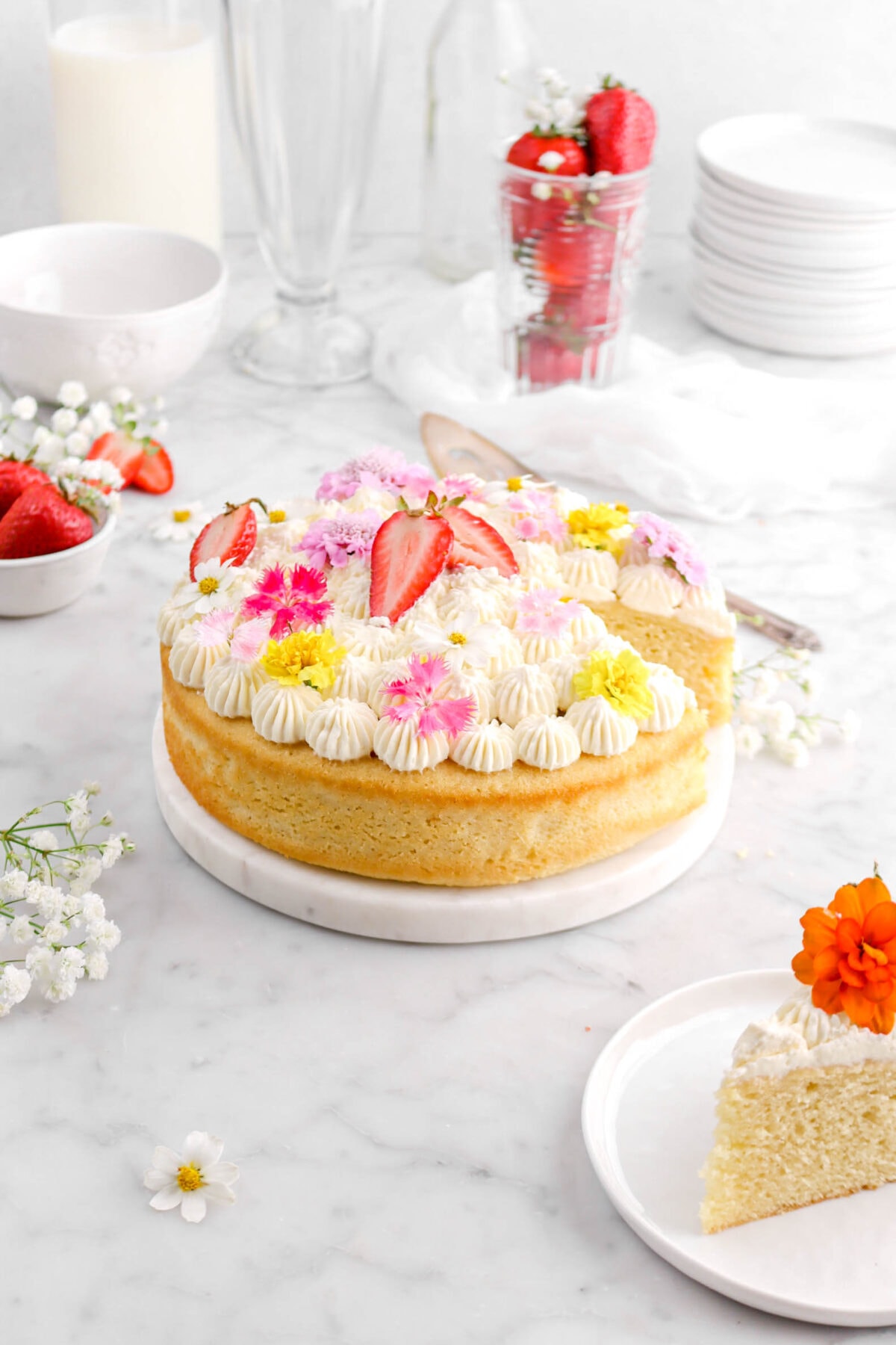 chamomile cake on marble plate on marble surface with piped frosting, strawberry halves, and flowers on top with slice of cake on white plate in front, flowers and strawberries around, and stack of white plates and glass of milk behind