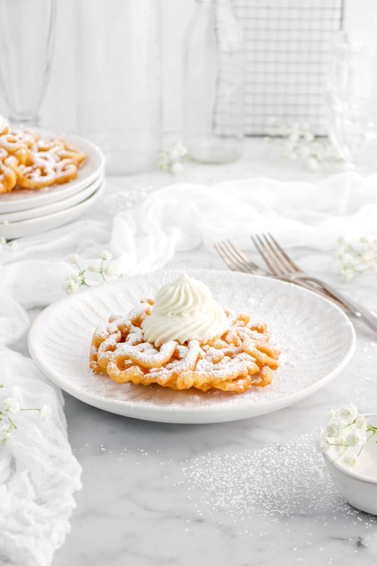 funnel cake on white plate with cheese cloth, forks, and flowers around