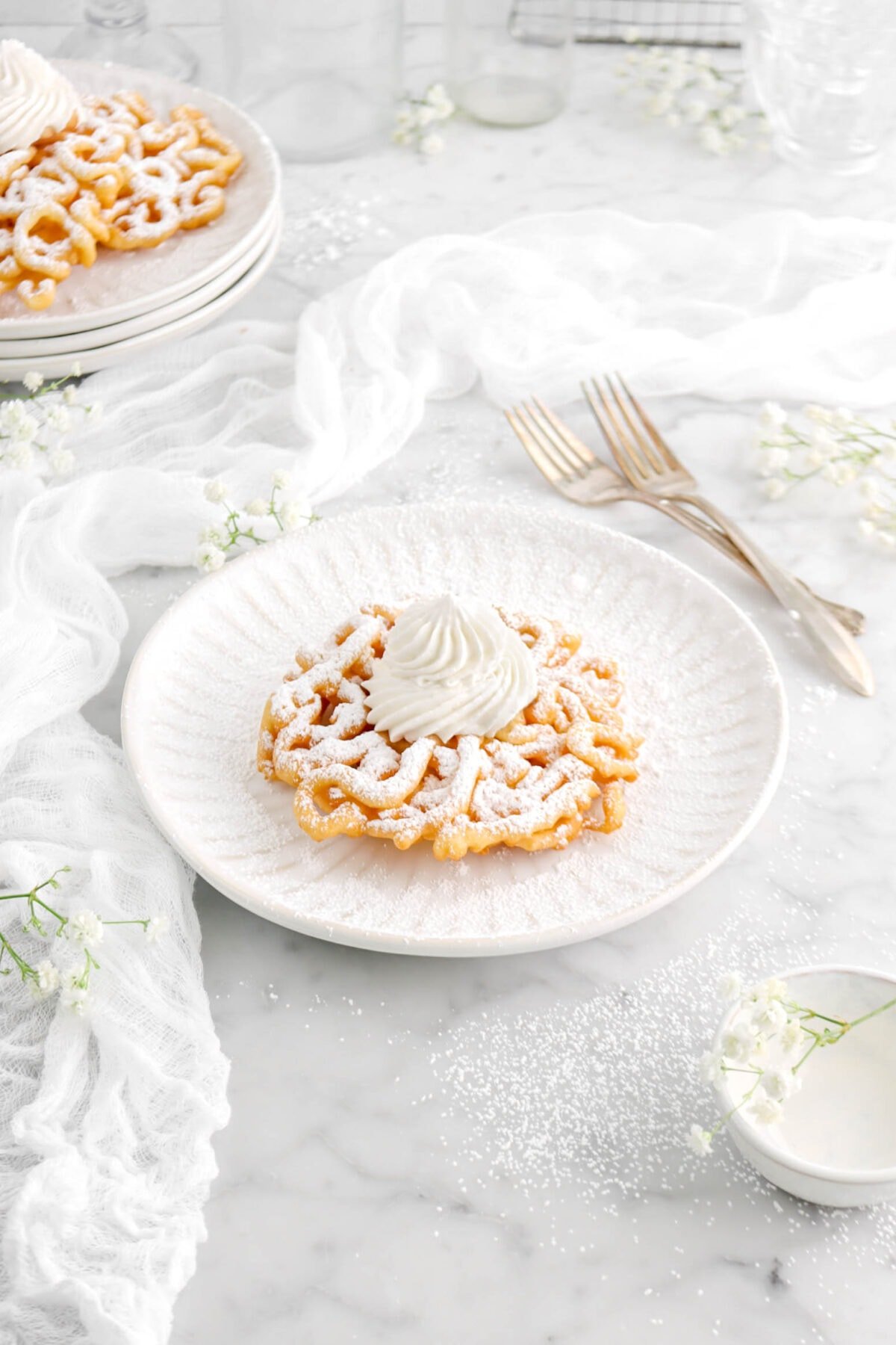 angled phto of funnel cake on white plate with powdered sugar and whipped cream on top, white cheese cloth behind, two forks, and flowers around