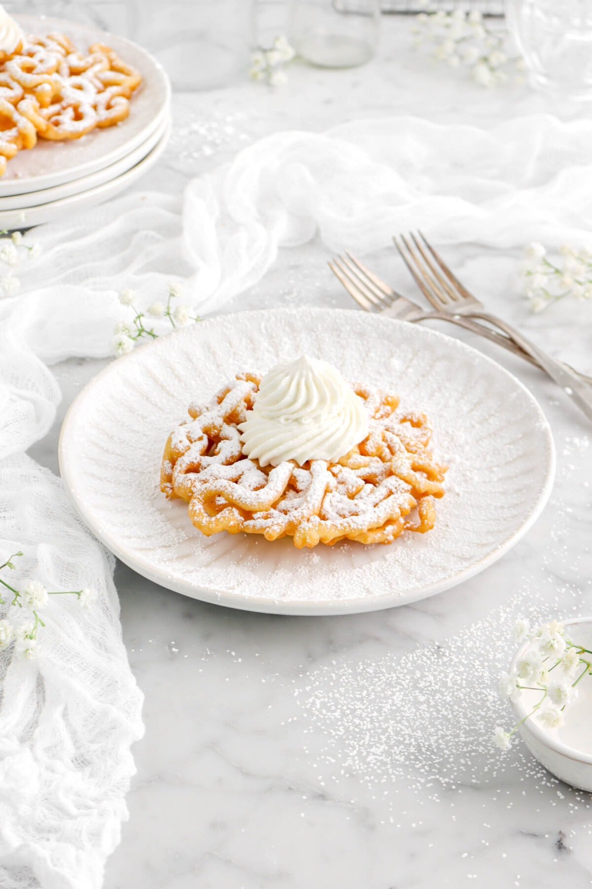 funnel cake with piped whipped cream and powdered sugar on top with white cheese cloth, flowers, and forks beside