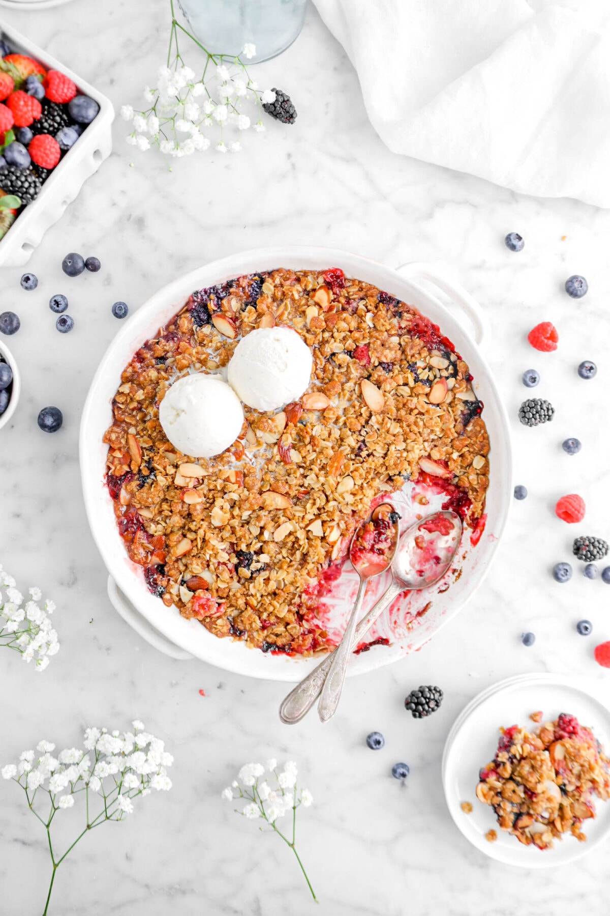 mixed berry crisp in white casserole with two spoons sitting inside, two scoops of ice cream on top, mixed berries around, and slice of crisp on stack of white plates beside