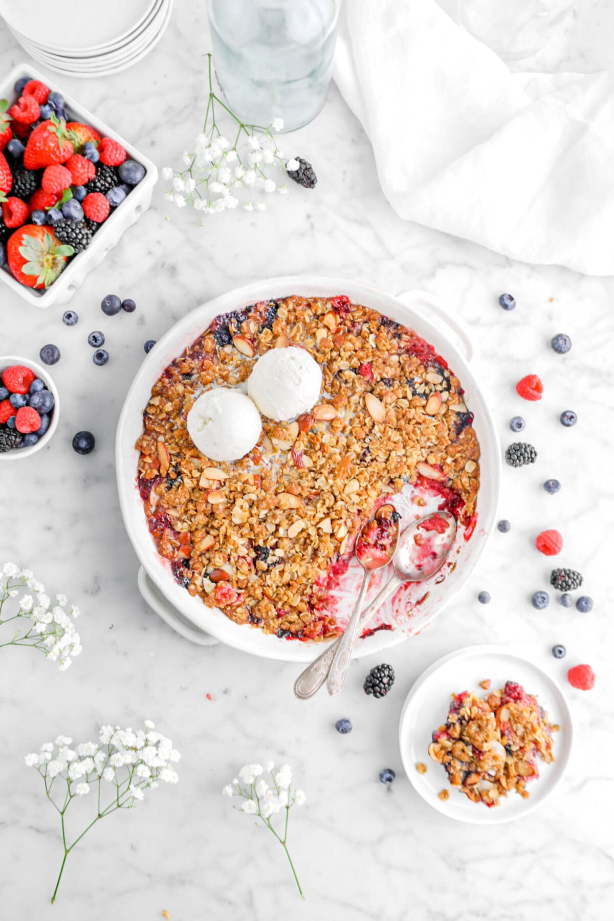 mixed berry crisp in white casserole with two scoops of ice cream on top, scoop of crisp on stack of white plates beside, and two spoons inside the crisp