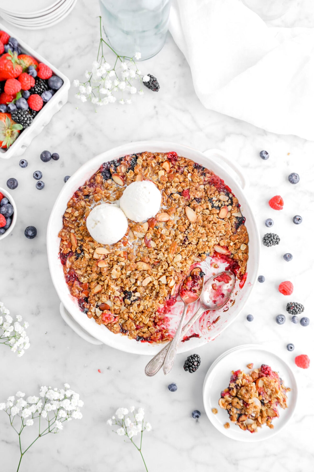 overhead shot of mixed berry crisp on marble surface with place of crisp beside, two spoons inside casserole, two scoops of ice cream on top of crisp, and mixed berries around
