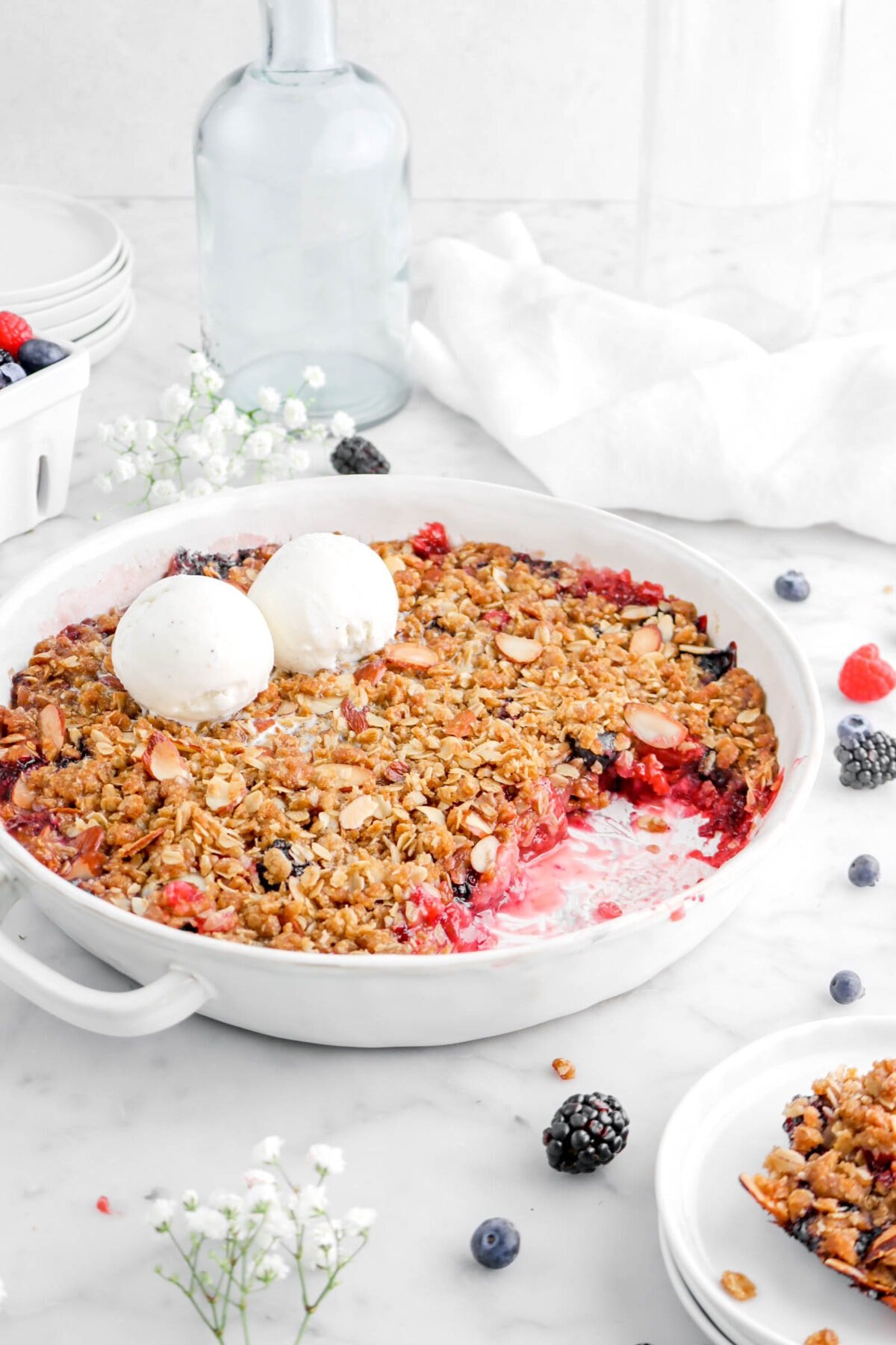 front shot of mixed berry crisp in round white casserole with two scoops of ice cream on top with empty glasses and white napkin behind