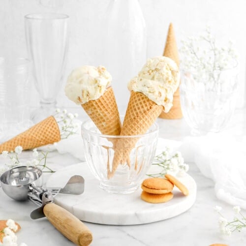 two sugar cones in glass bowl with two scoops of ice cream on right cone and one scoop of ice cream on left cone, antique ice cream scoop beside, more sugar cones behind, and vanilla wafers around