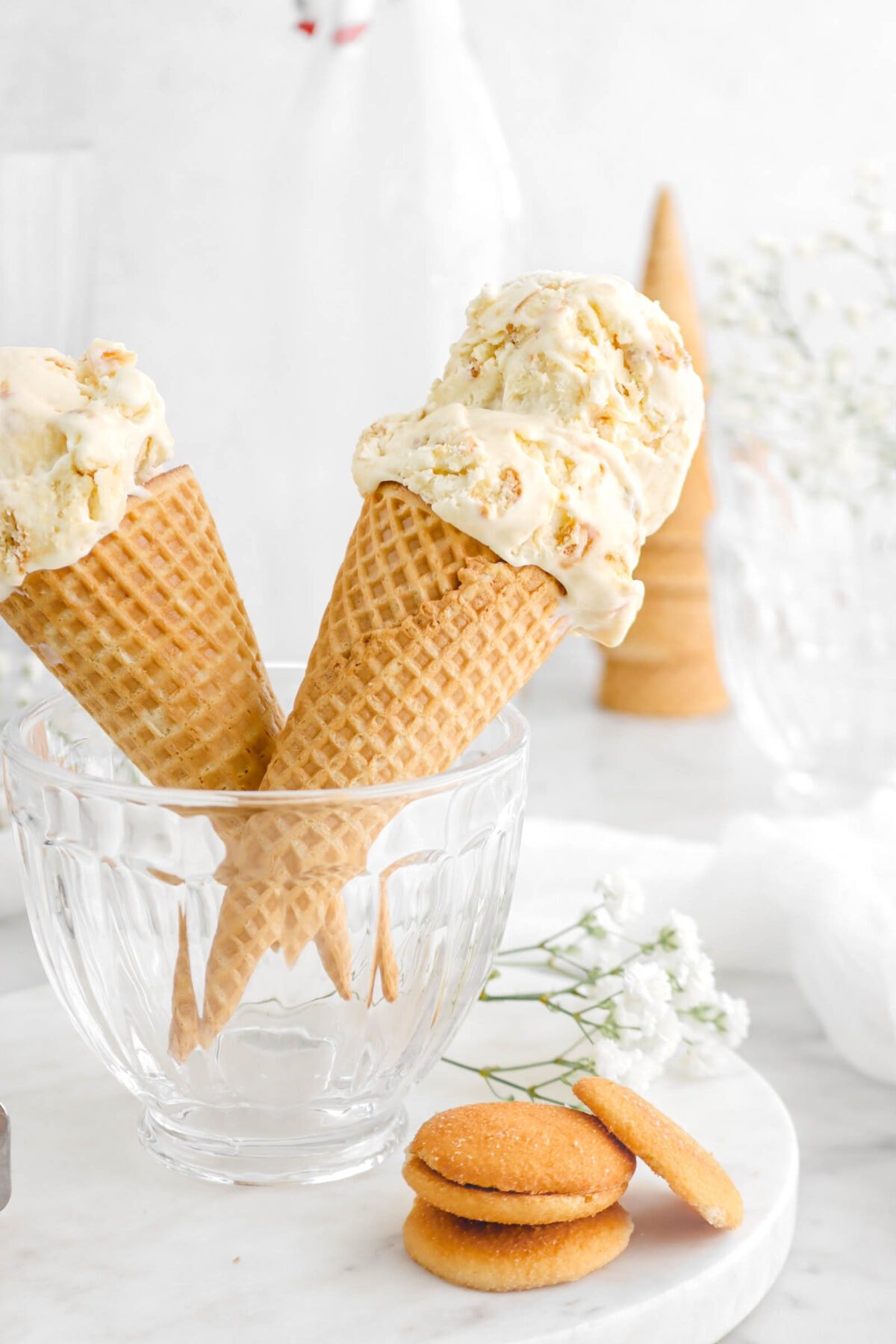 close up of banana pudding ice cream in a sugar cone in a glass bowl with vanilla wafers and flowers beside