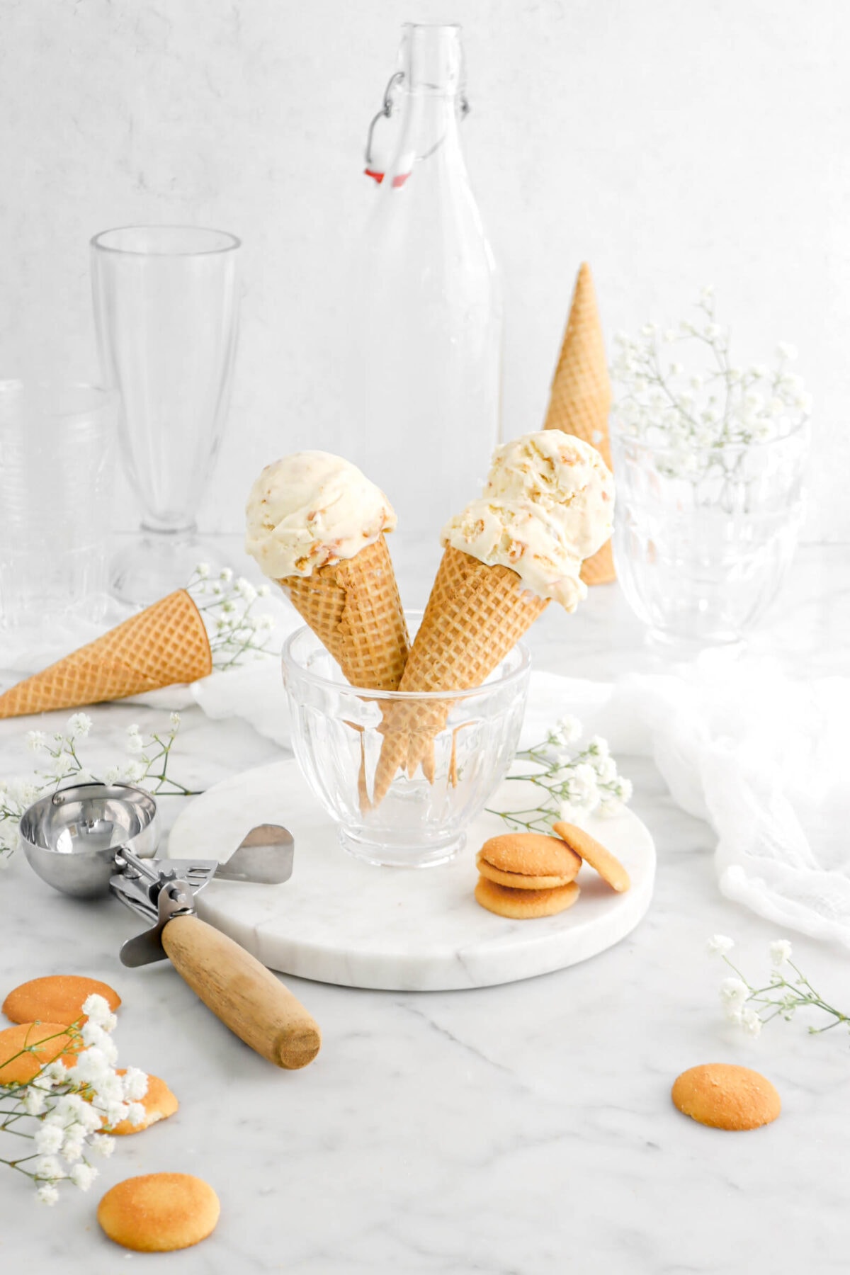 pulled back shot of banana pudding ice cream in two sugar cones in a glass bowl on a marble plate with empty cones behind, vanilla wafers and flowers around, an antique ice cream scoop beside, and an empty milkshake glass behind